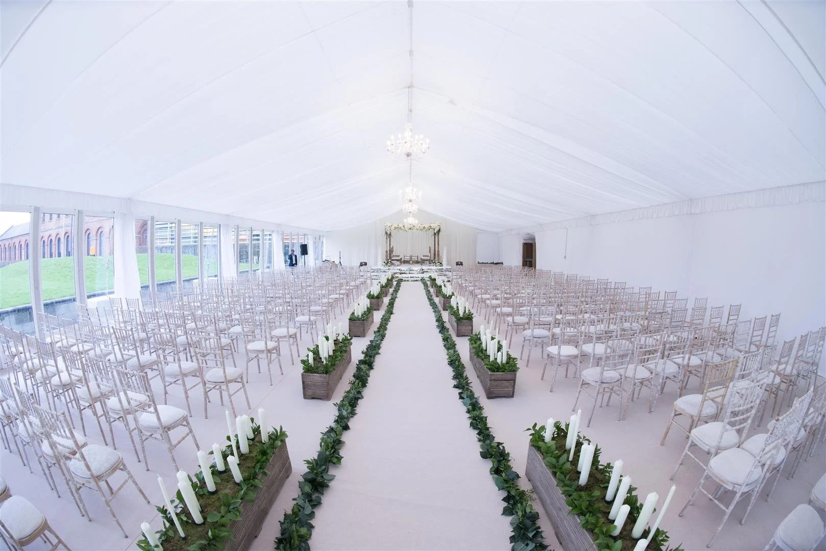 White wedding ceremony setup inside a large tent with rows of white chairs, a central aisle decorated with green foliage and candles in rectangular pots, a floral arch and chandelier lighting at the front, and windows along one side letting in natural light.