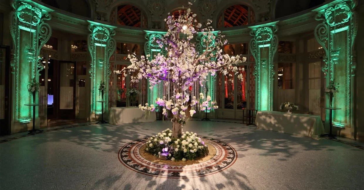 Elegant indoor venue with a central decorative lit cherry blossom tree surrounded by white flowers, ornate architecture with green lighting accents, and draped tables along the walls.