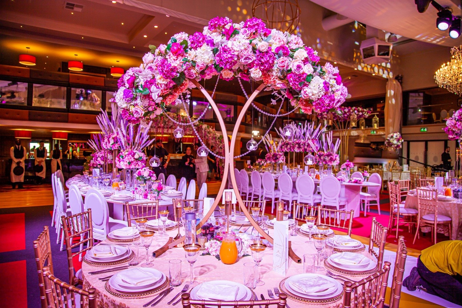 A banquet hall decorated with pink and purple floral centerpieces, including a large floral tree centerpiece atop a round table, and elegant dining settings with plates, glasses, and cutlery. The hall features warm lighting, chandeliers, and multiple decorated tables with chairs, suited for a celebration or wedding reception.