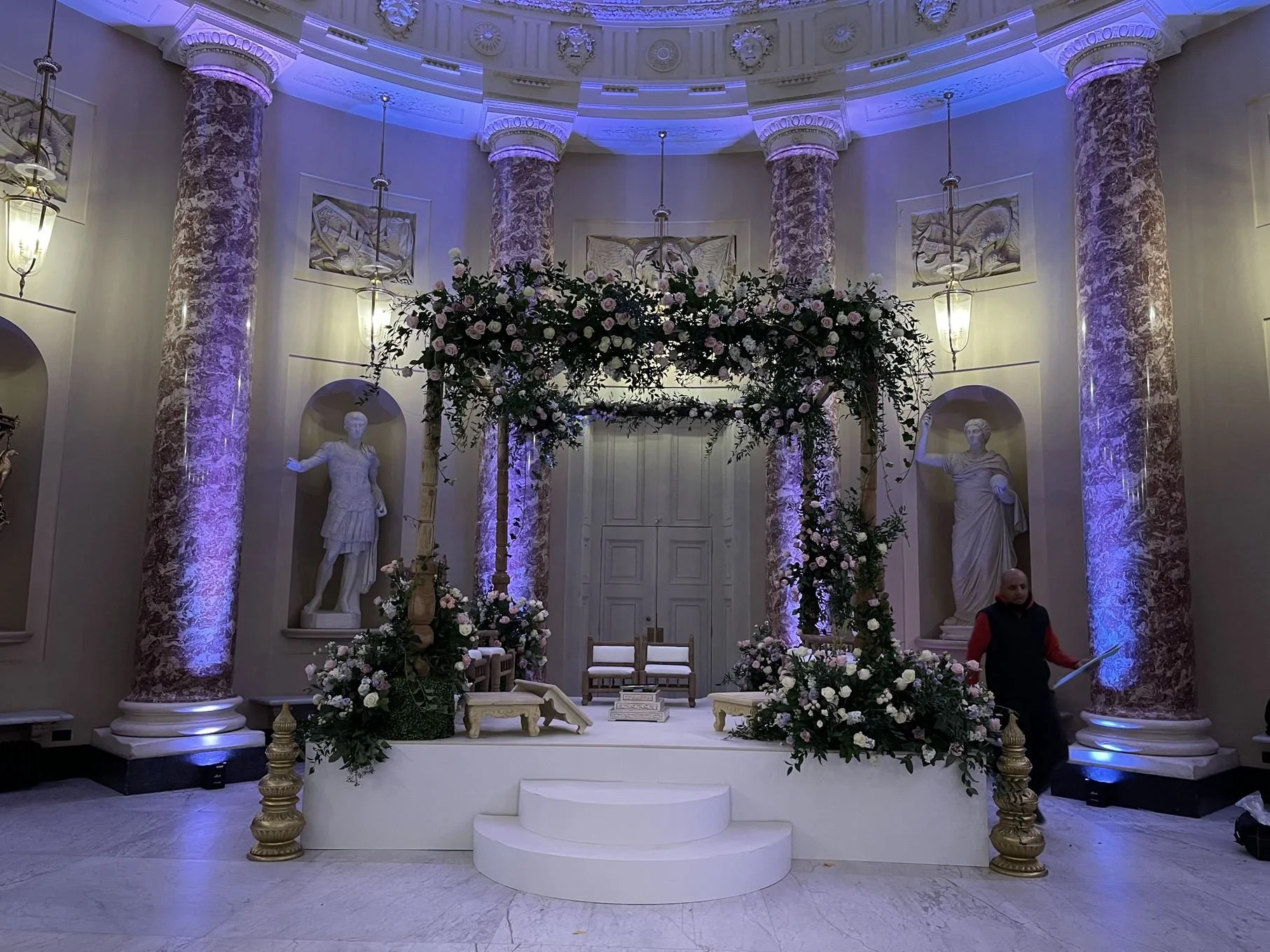 A decorated wedding or event stage with marble columns, floral arrangements, and classical statues, illuminated with purple and blue lighting.