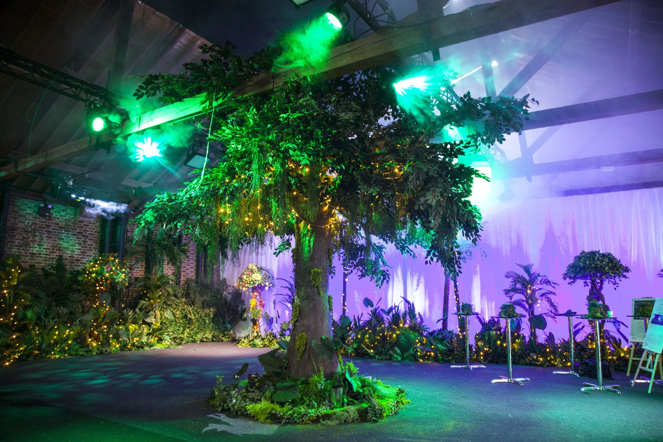 Indoor scene with a large artificial tree decorated with string lights, surrounded by tropical plants, colorful lighting, and a purple-lit backdrop.