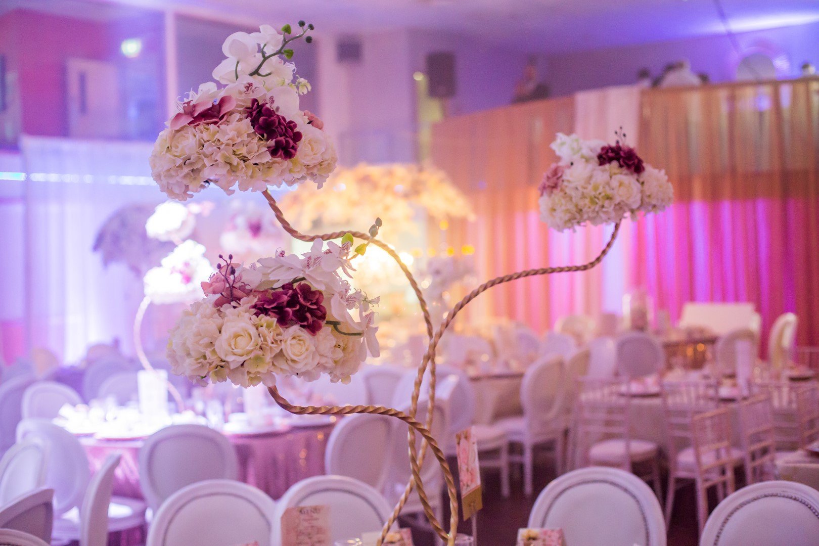 Elegant event venue decorated with floral centerpieces, featuring white and purple flowers on twisted branches, pink lighting, and round tables with white chairs.