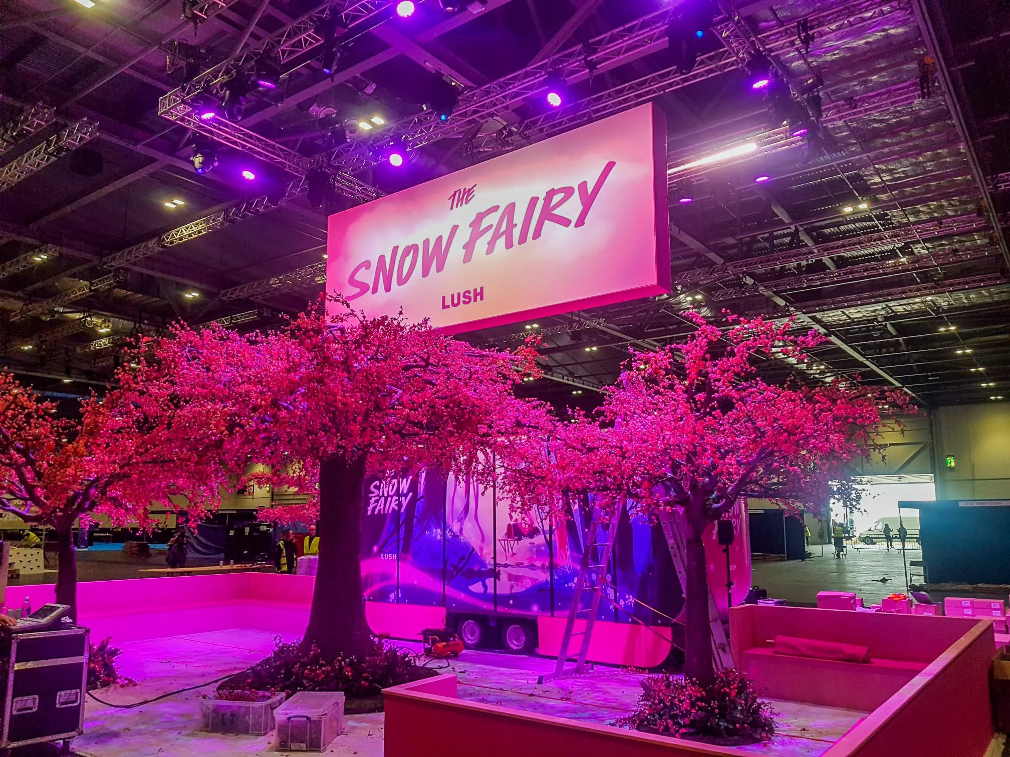 Trade show booth decorated with pink artificial cherry blossom trees and a large sign that reads 'The Snow Fairy Lush' in pink and purple font.