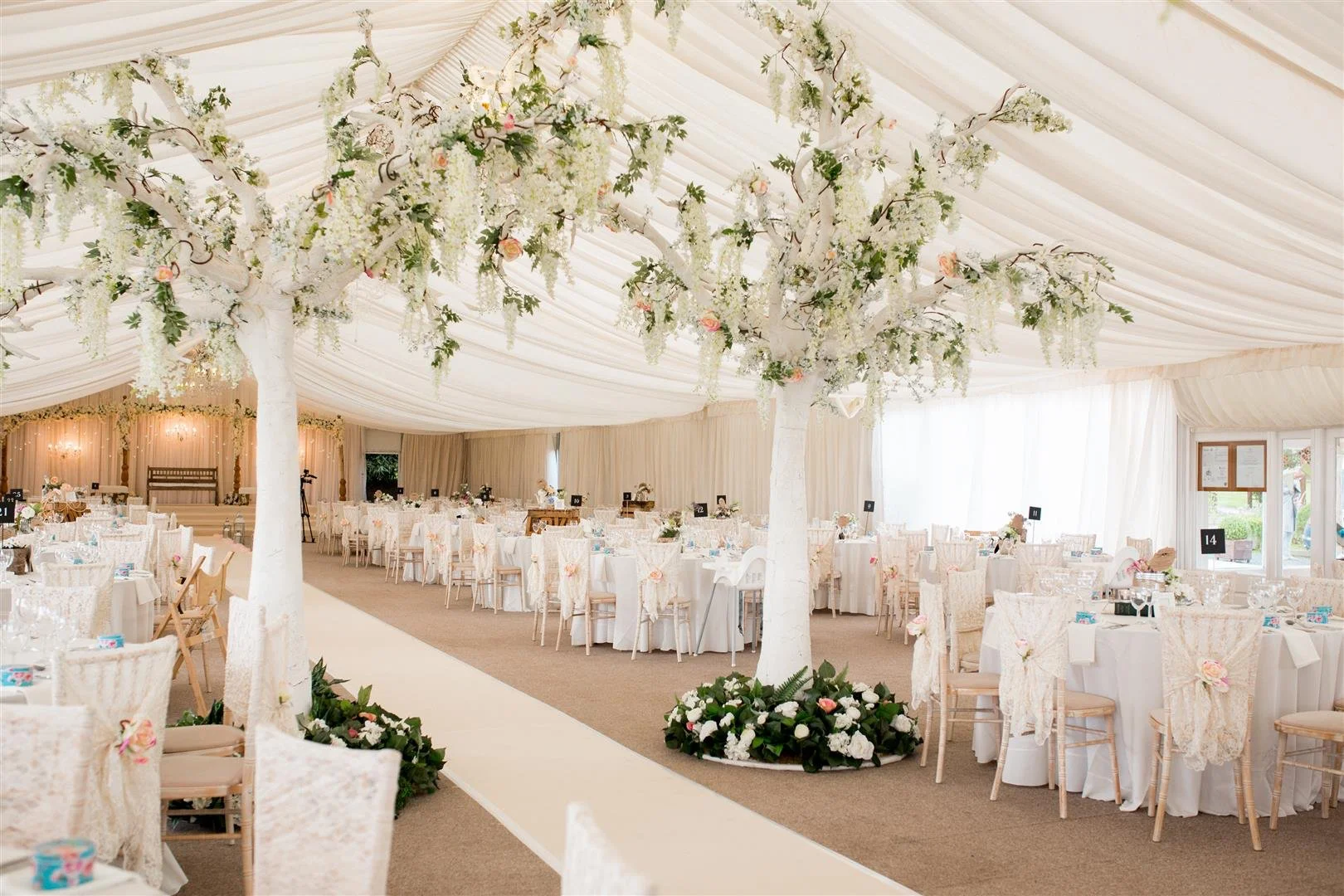 Elegant wedding reception tent decorated with large white floral trees, round tables with white tablecloths, and pastel floral arrangements, illuminated with soft lighting.