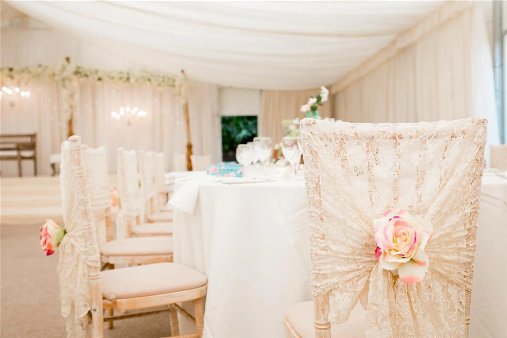Decorated wedding reception chairs with lace covers and pink roses, set in a bright, elegant tent with a decorated table and floral backdrop.