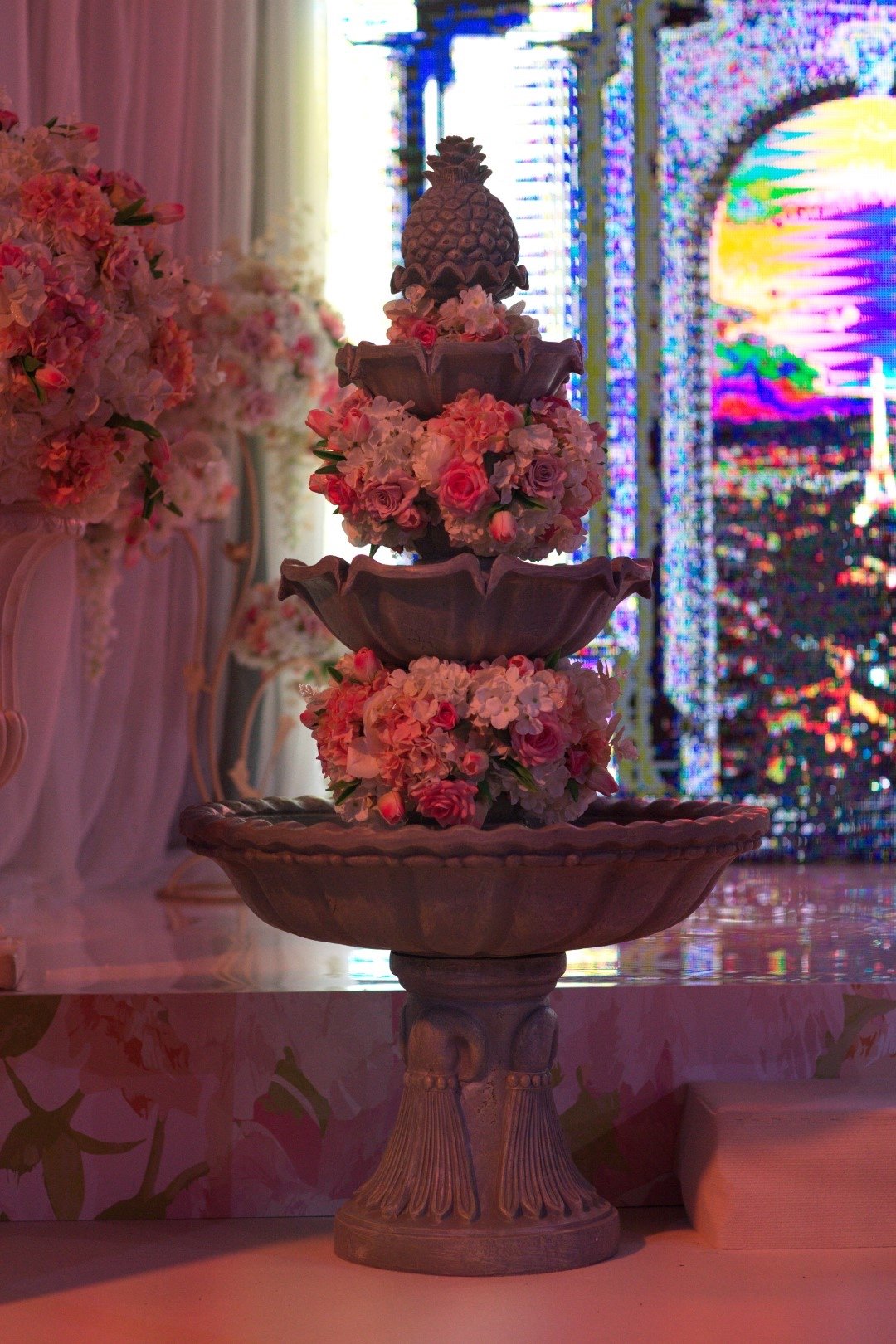 A multi-tiered fountain decorated with pink and white flowers, with a pineapple finial on top, situated in a decorated setting with pink floral arrangements and digital screens in the background.