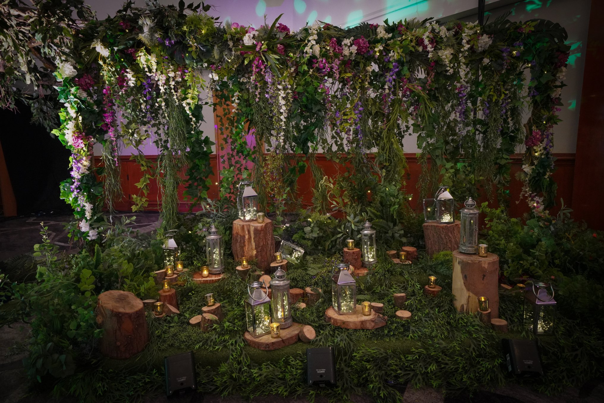 A decorative display with hanging greenery and purple and white flowers, wooden stumps, and lanterns with lights, arranged on a lush green base for an event or celebration.