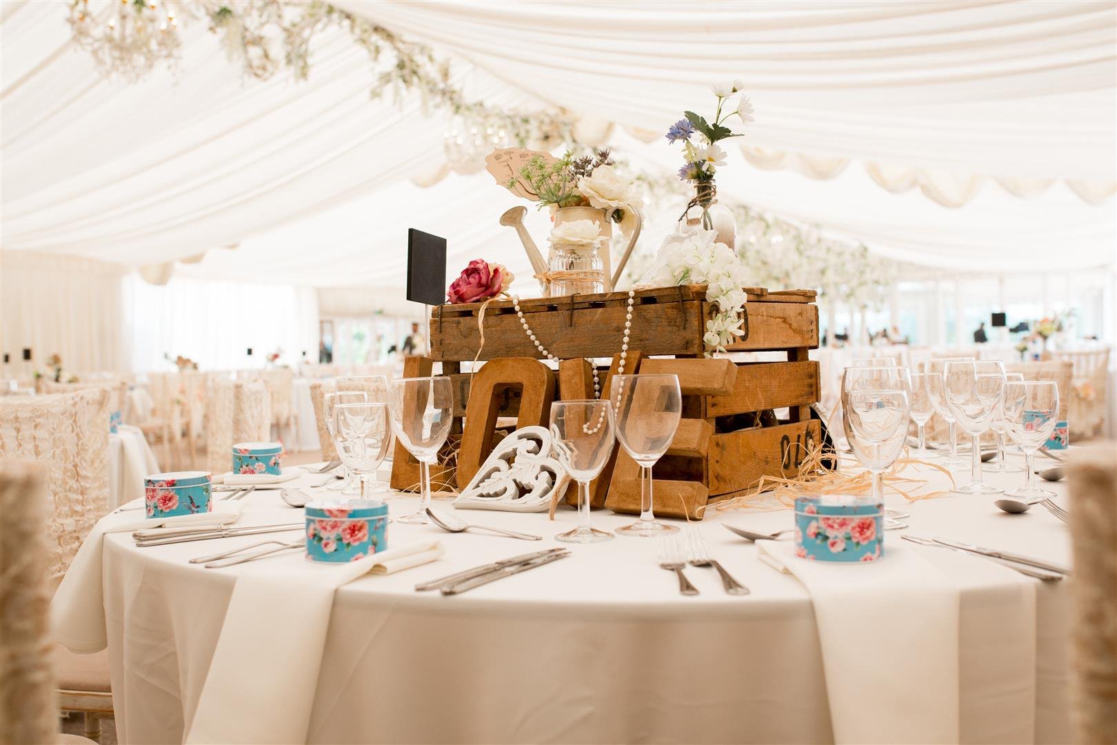 Wedding reception table with a wooden centerpiece decorated with flowers, beads, and small chalkboard signs, set with glassware, silverware, and colorful boxes, under a white draped tent.