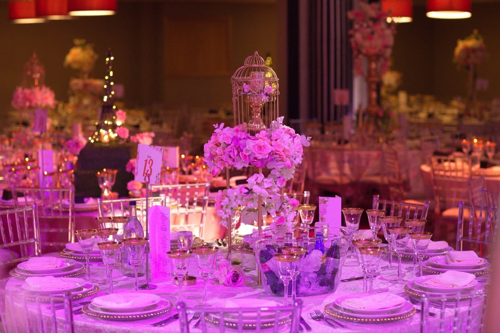 Elegant banquet table with pink and purple floral centerpiece, glassware, plates, and table number card, set in a decorated event hall.