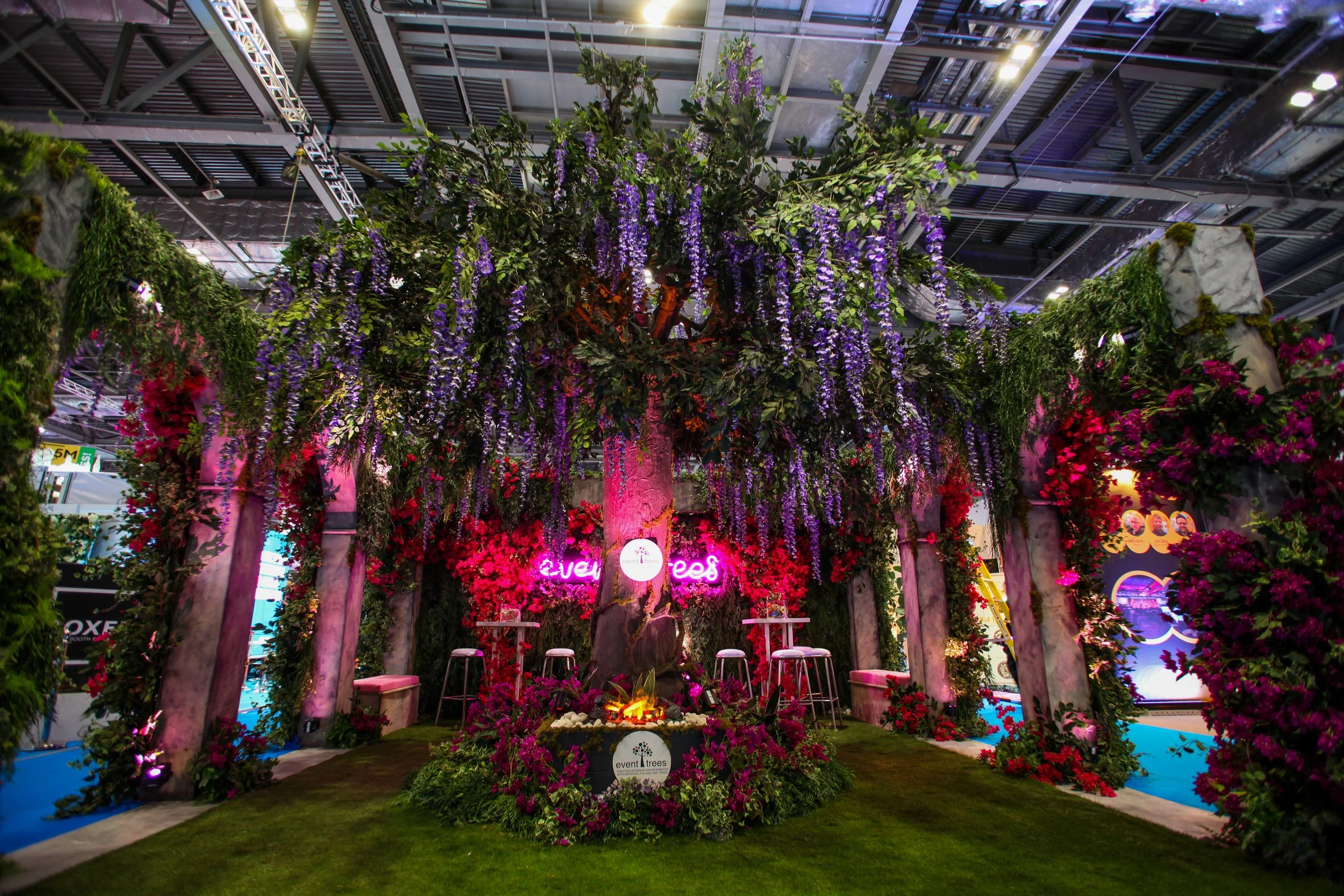 Indoor garden display with purple and pink flowers, green foliage, wooden pillars, and a neon sign that reads 'event trees' inside a building with a high ceiling and metal framework.