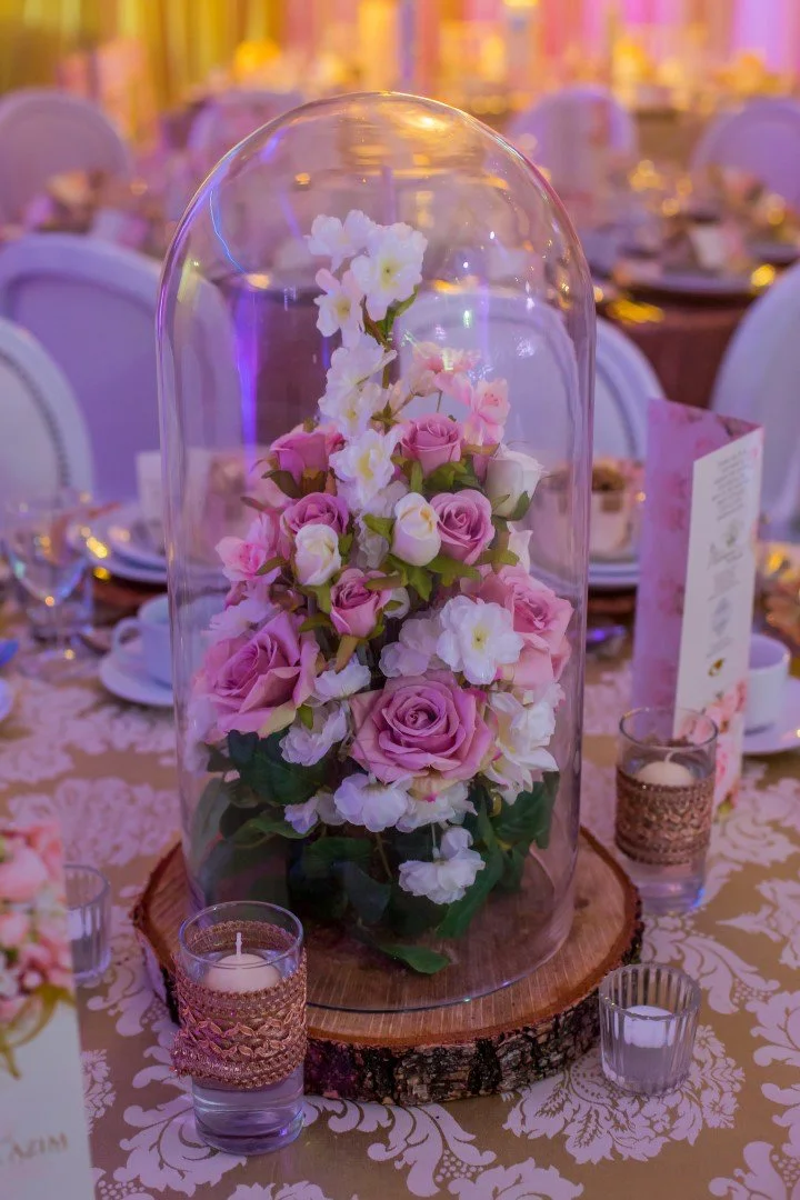 A floral centerpiece with pink roses and white flowers inside a glass dome on a wood base, surrounded by small lit candles, at an elegant event with decorated tables.