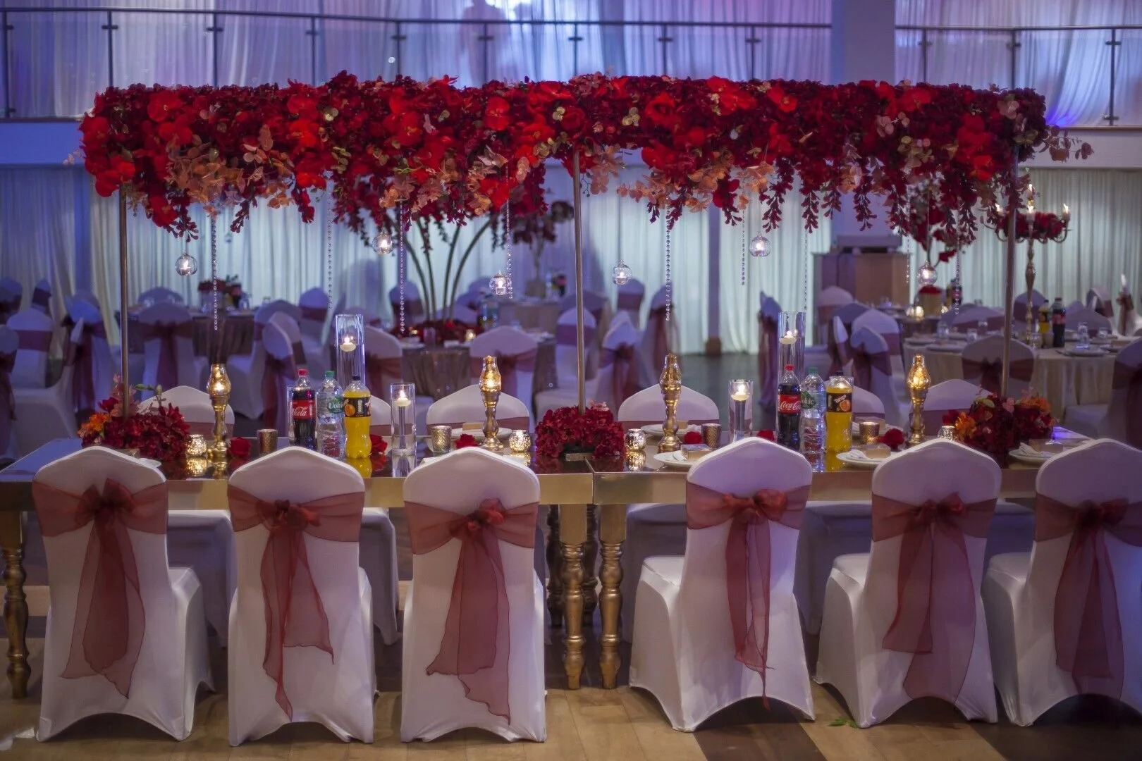 Decorated banquet hall with pink and red floral centerpieces, white chairs with pink bows, and candles on tables.