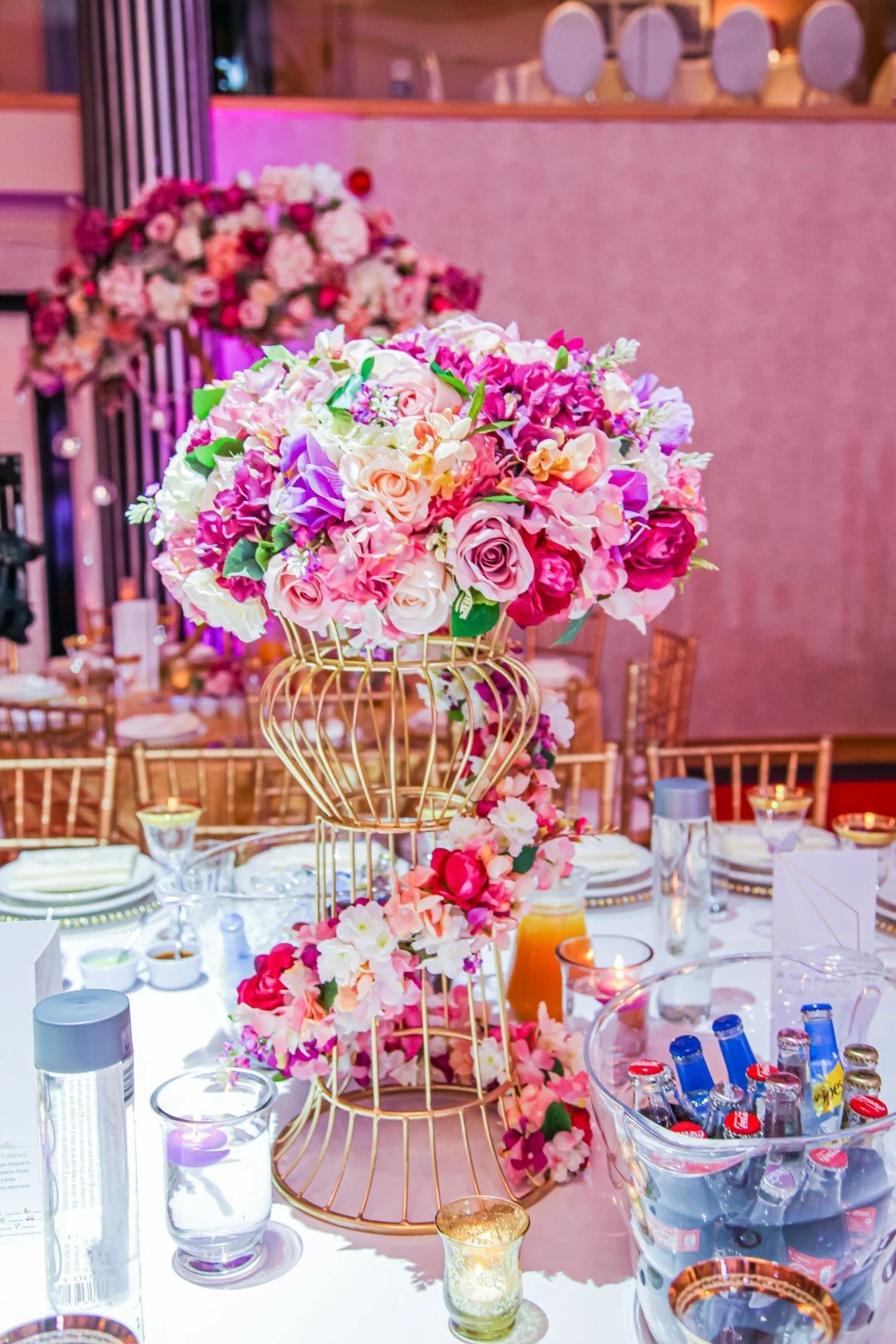A floral centerpiece with pink and white roses, orchids, and other flowers in a gold wire cage on a decorated table at an event.