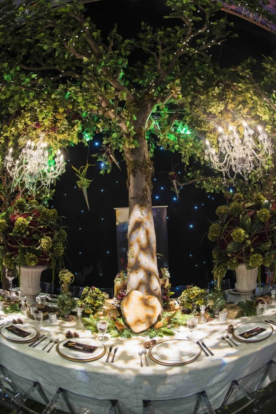 A decorated banquet table with a large illuminated tree centerpiece, chandeliers, flowers, and elegant table settings for a festive event.