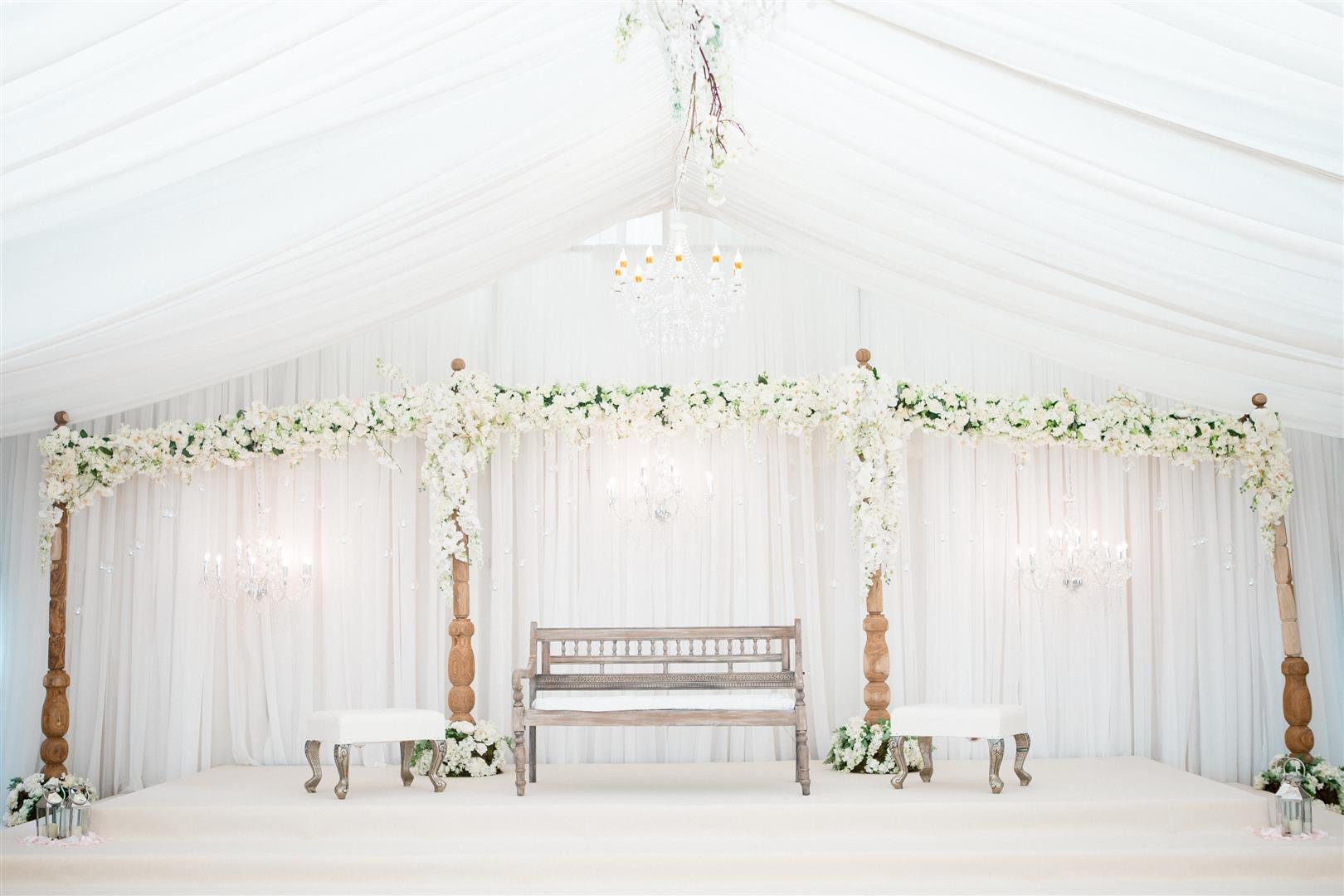Elegant wedding stage decorated with white flowers, drapery, chandeliers, and wooden furniture.