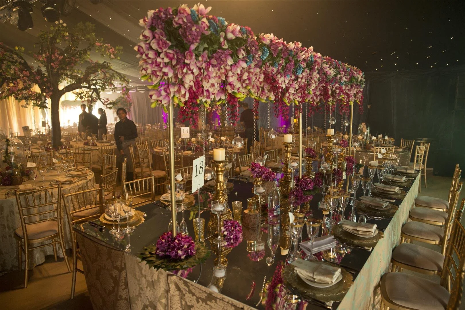 Elegant banquet table decorated with tall floral centerpieces of pink and purple flowers, surrounded by gold chairs and set with plates, napkins, and glassware, in a warmly lit event hall.