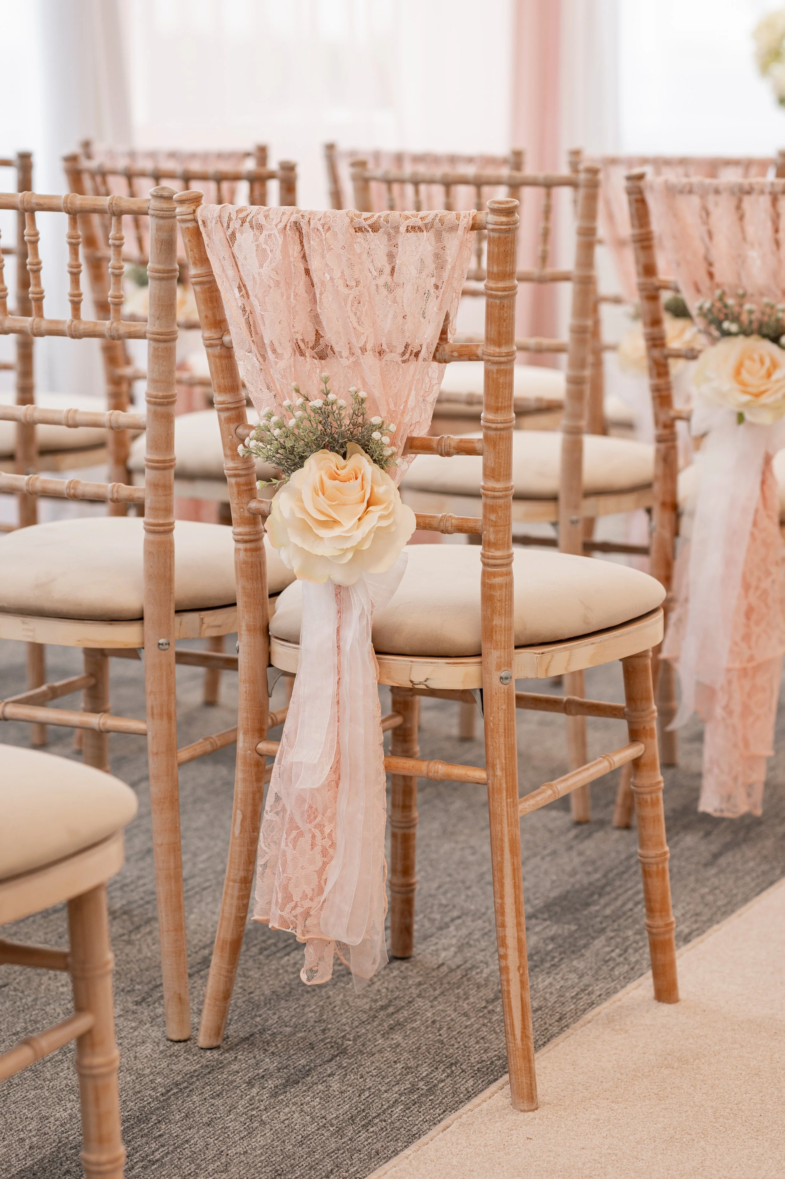 Decorated wooden chairs for a wedding ceremony, featuring lace ribbons and floral arrangements with roses and small white flowers.