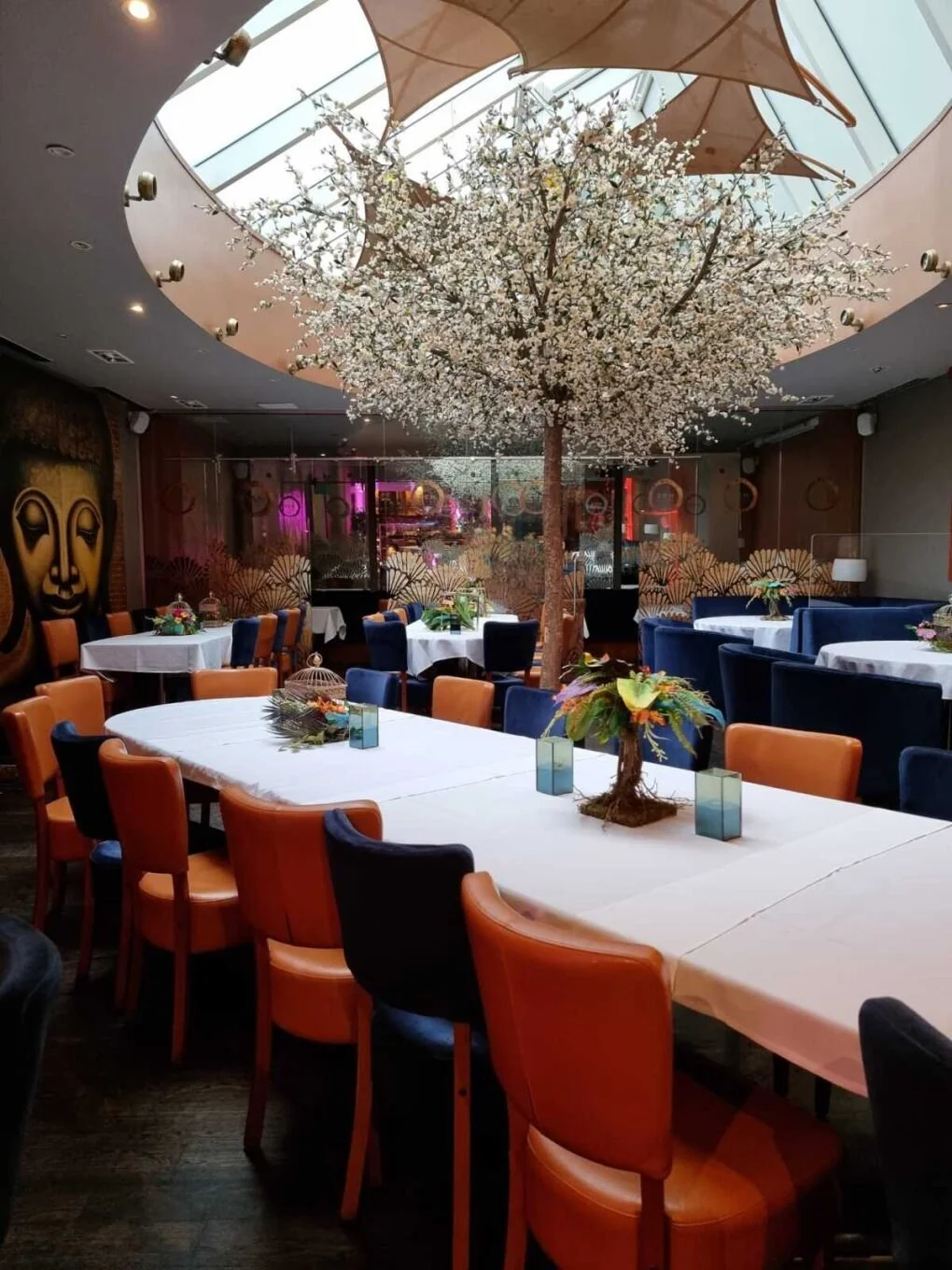 Indoor restaurant with a large cherry blossom tree centerpiece, surrounded by white tablecloths and colorful chairs, and a Buddha wall art in the background.