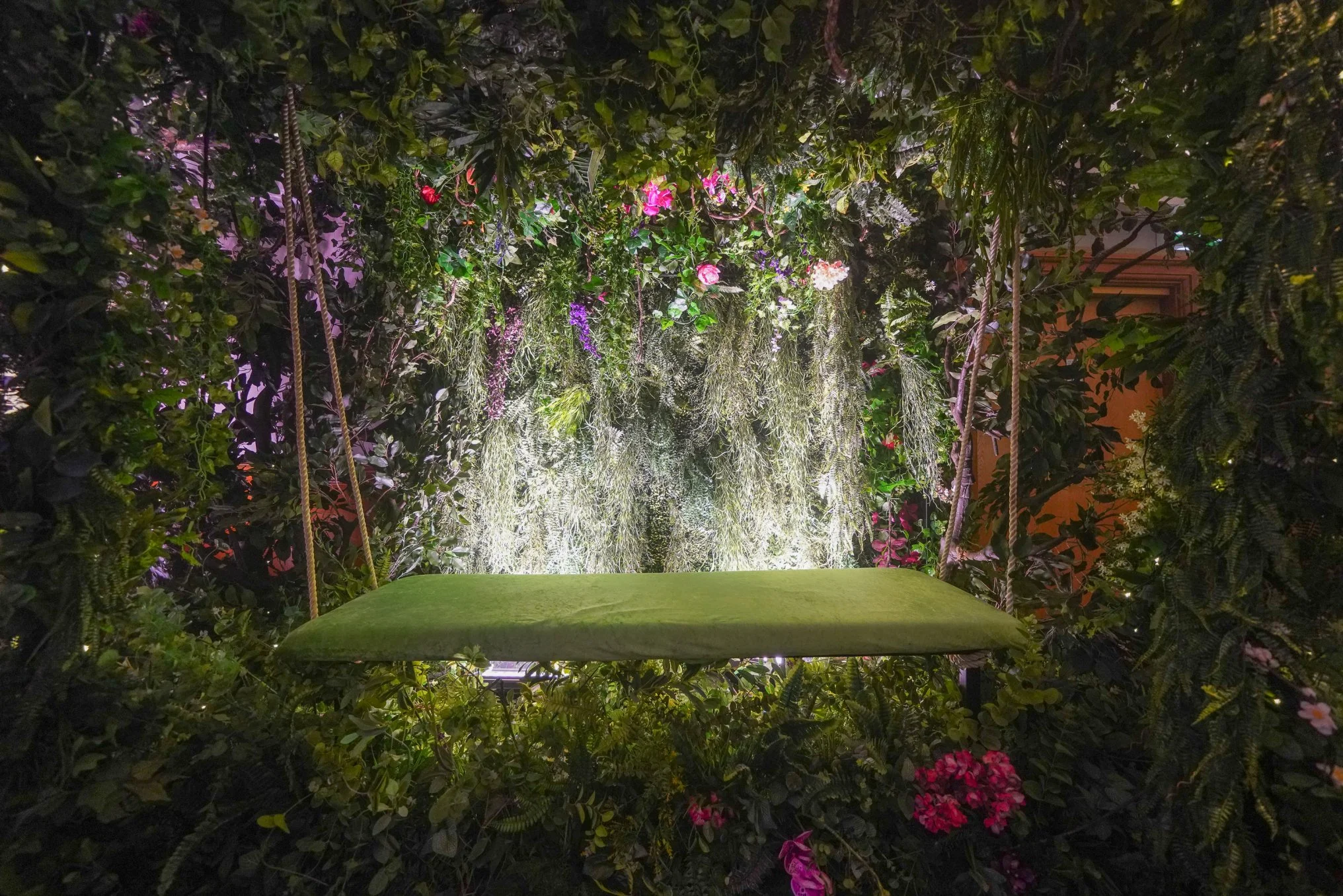 Green swing seat hanging amidst lush green garden walls and hanging plants, with soft lighting and pink and purple flowers.