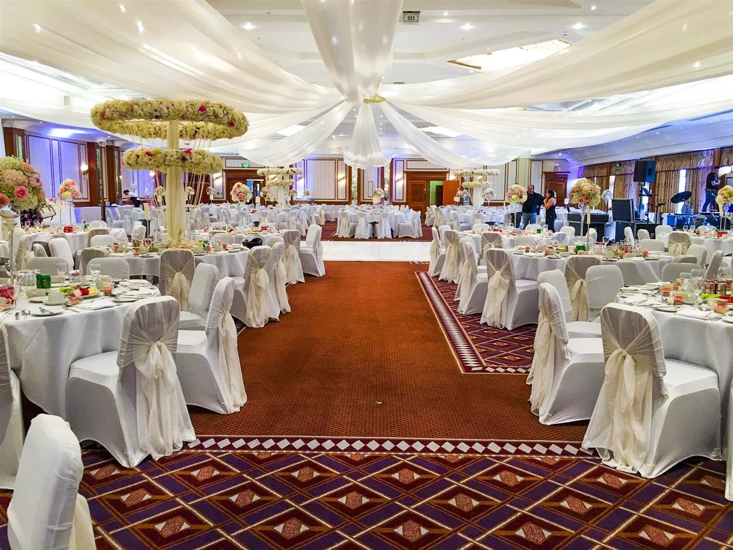 Elegant banquet hall decorated for a wedding reception with round tables covered in white tablecloths, decorated with floral centerpieces, and chairs with white covers and bows. The ceiling has draped white fabric, and there are floral arrangements on tall stands. The room has patterned carpet, soft lighting, and a stage area with musical instruments.