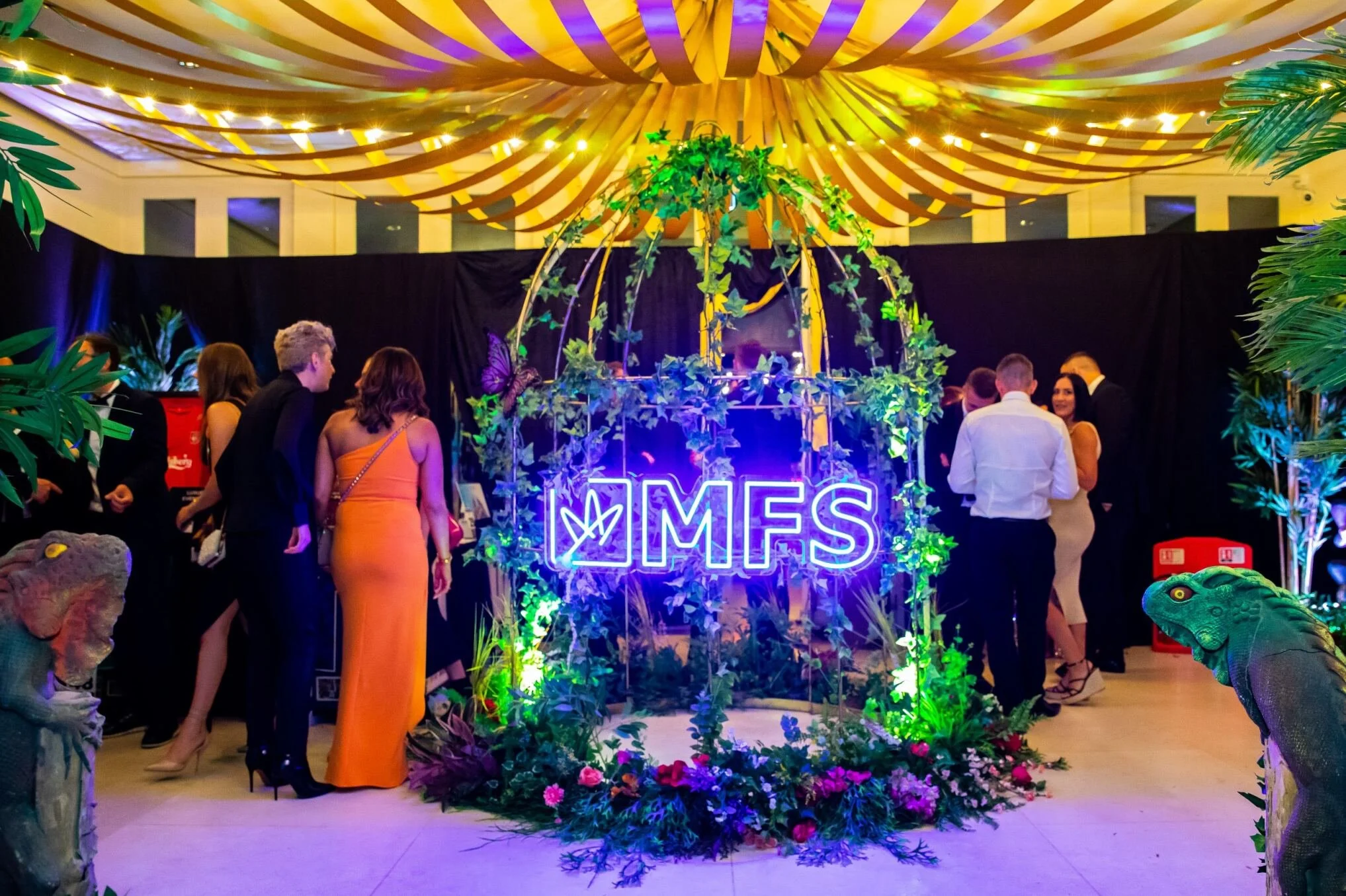 An indoor event featuring tropical theme decorations with lush greenery, giant animal sculptures, and a neon sign reading 'MFS' surrounded by flowers, with guests dressed in formal attire.