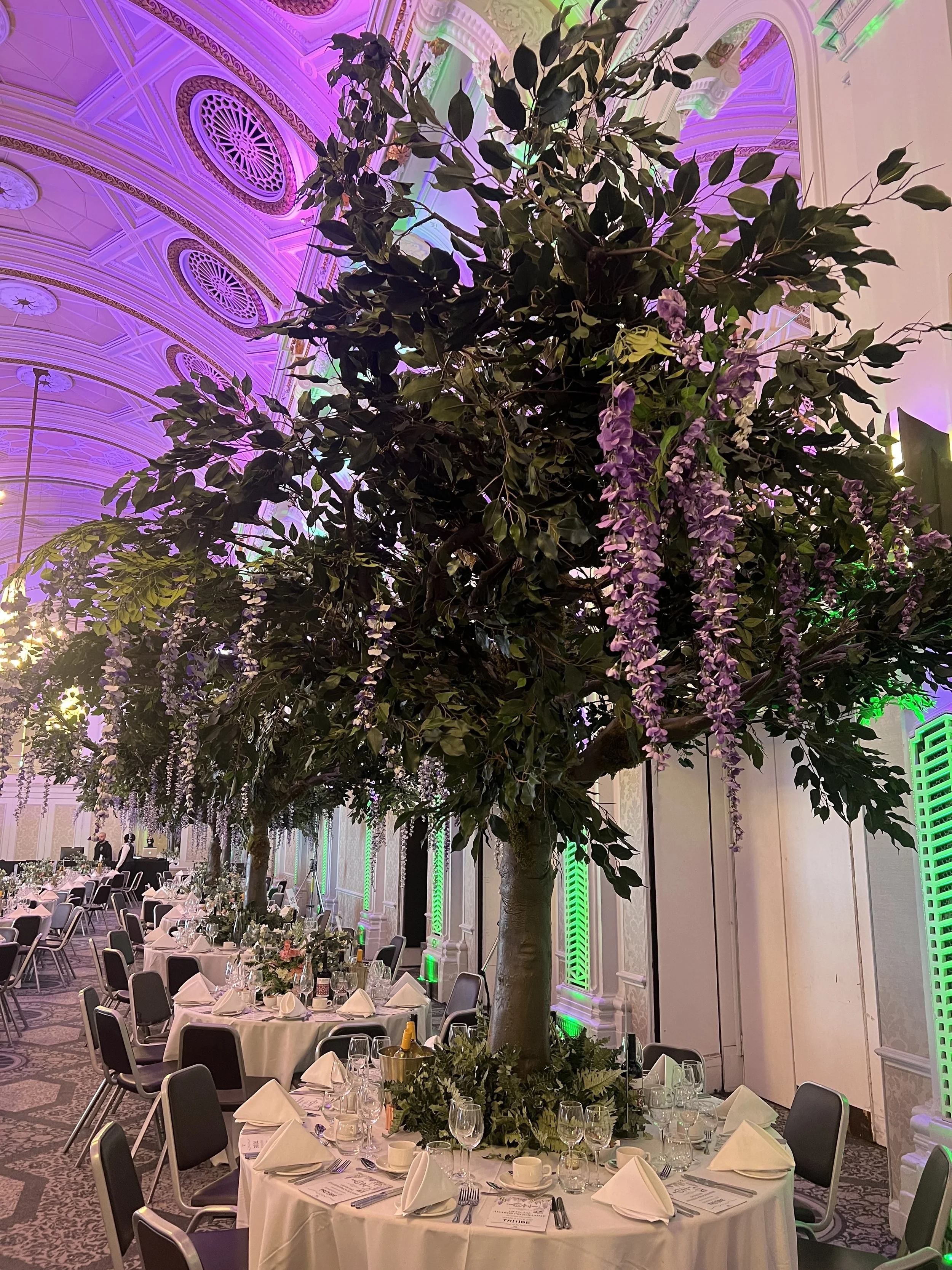 Decorated banquet hall with a large artificial tree with purple hanging flowers centered on a round table set for a formal event with white tablecloths, napkins, glassware, and plates, under an ornately decorated ceiling with purple and green lighting.