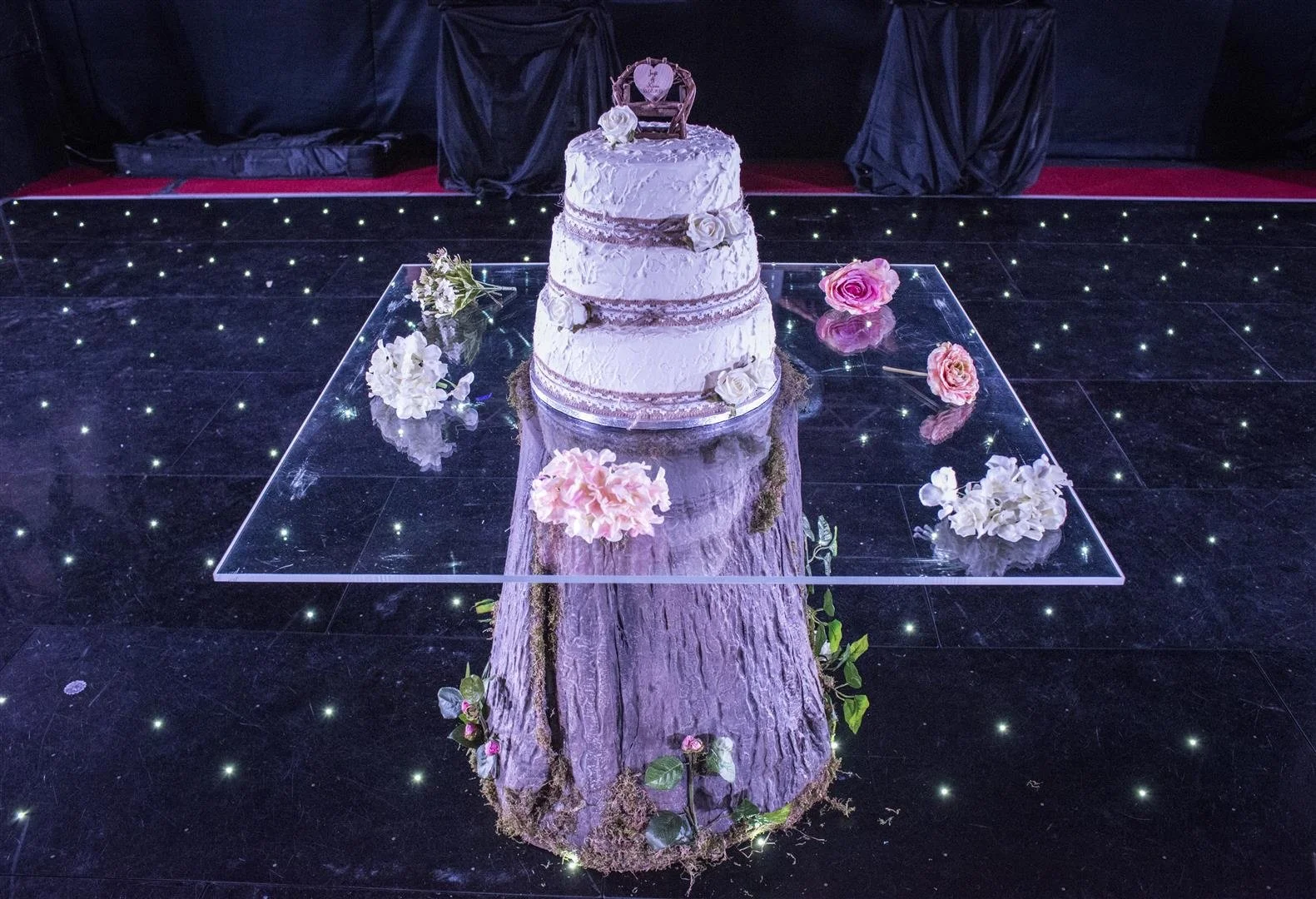 A white, layered wedding cake on a tree stump with floral decorations, placed on top of a glass platform on a black floor with small star-like lights. Flowers surround the platform, and a dark backdrop is in the background.