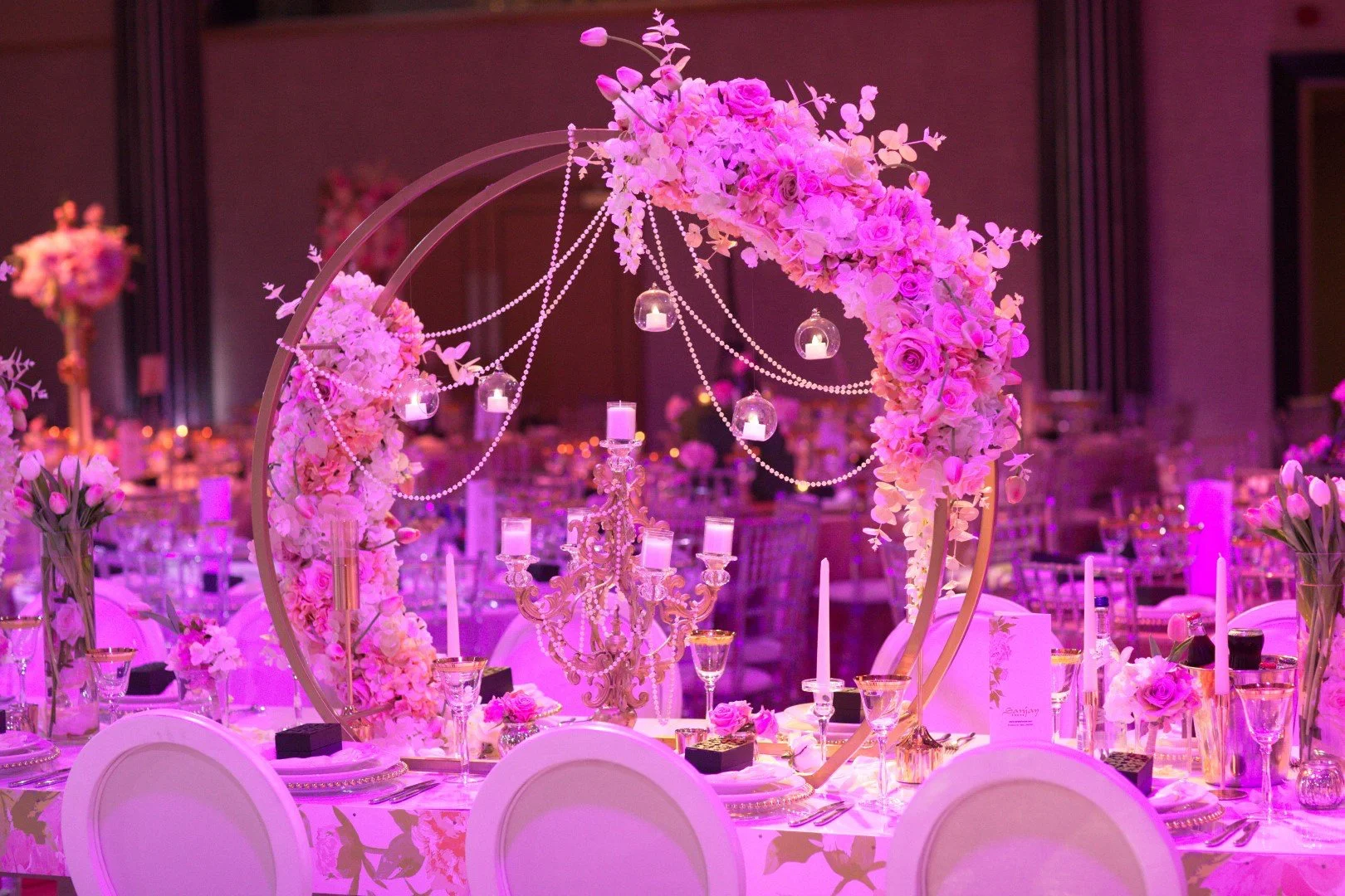 A decorated banquet table with a floral arch, pink and purple flowers, candles, and elegant glassware at a wedding or event.