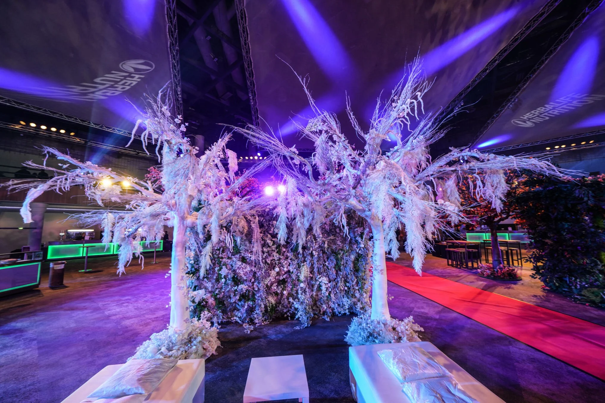 Decorative event space featuring white trees with branches covered in snow-like material, purple and blue lighting, and floral arrangements along a red carpet pathway.
