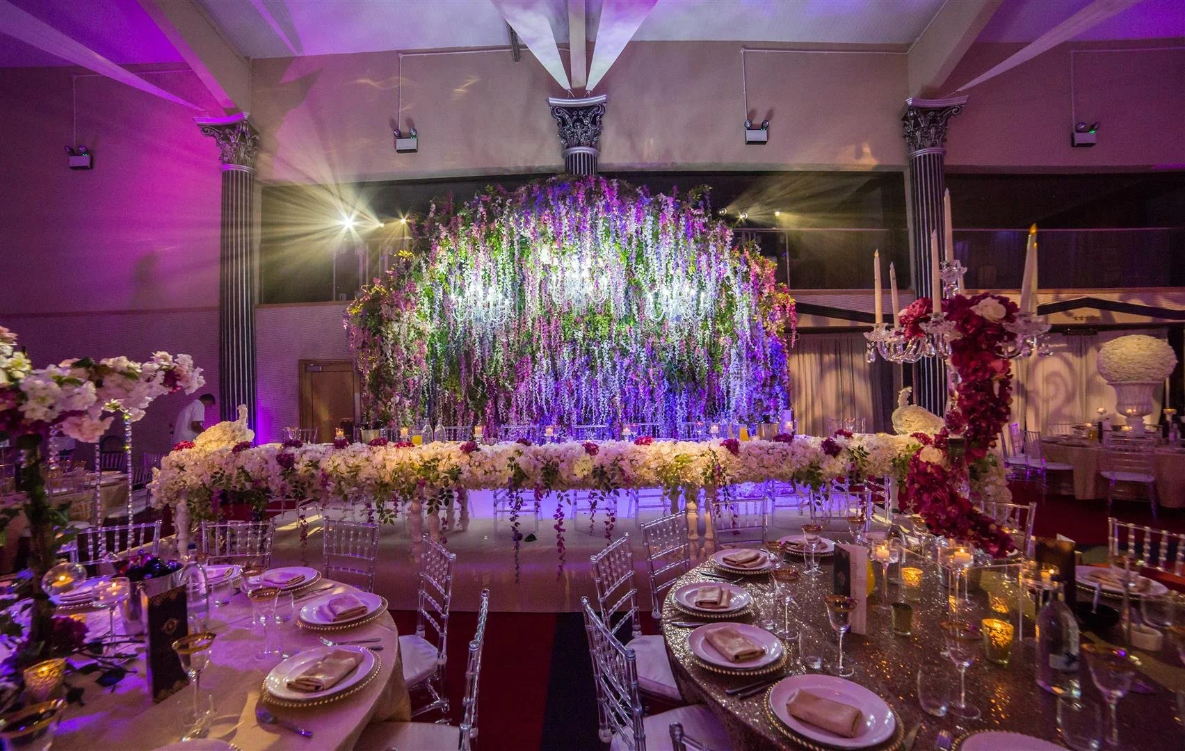 Elegant wedding reception setup with tables, floral arrangements, and a grand floral backdrop illuminated with purple lights.
