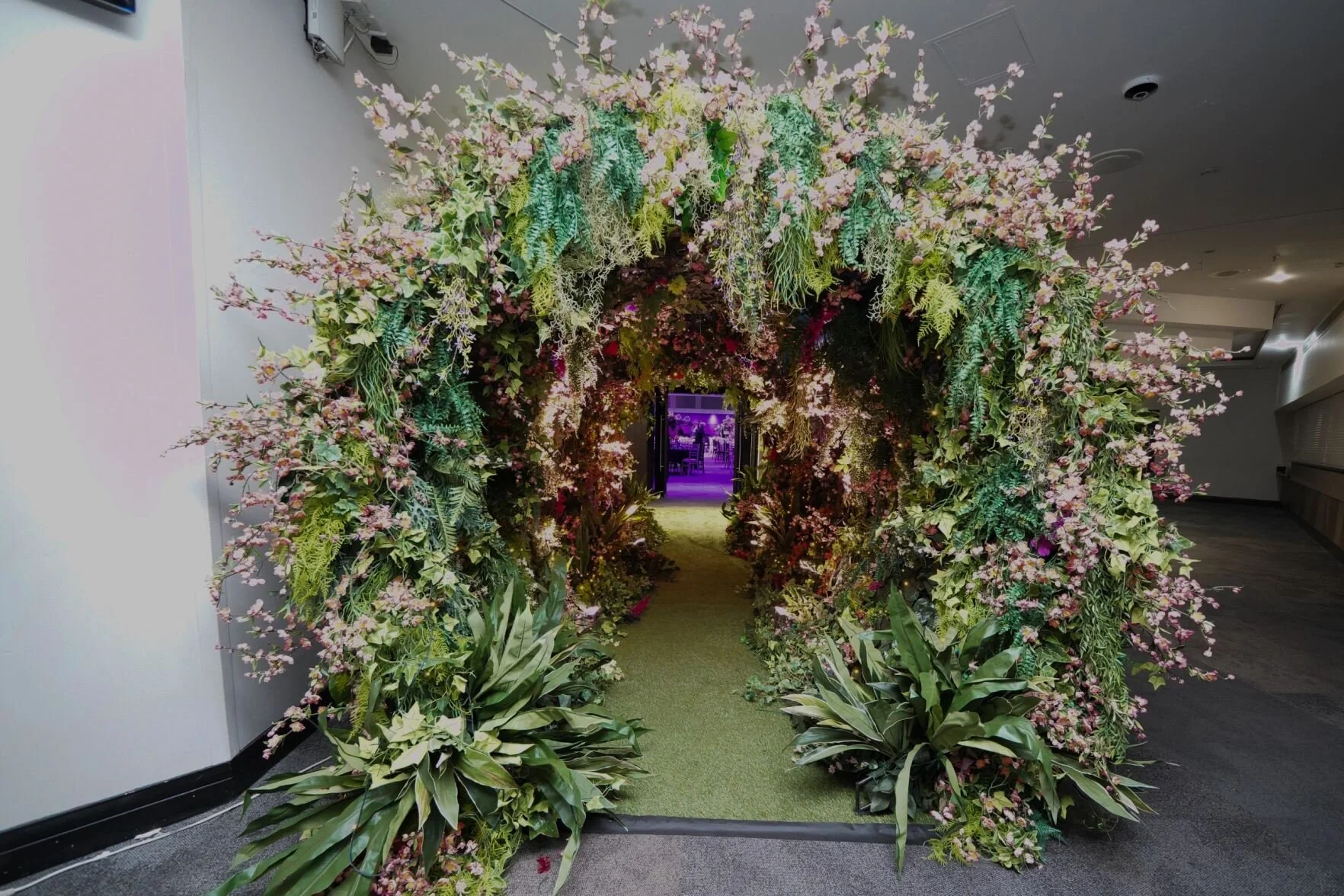 A floral archway decorated with pink and white flowers and green foliage leading to a hallway with purple lighting.