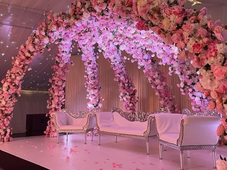 Elegant stage decorated with pink and white roses, featuring a floral arch and vintage-style white sofas and chairs for a wedding or special event.