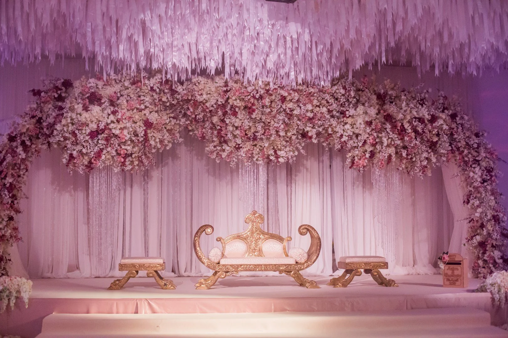 Elegant wedding stage decorated with pink and white flowers, draped pink fabric, and a purple chandelier overhead. There is a gold ornate throne with two matching benches on either side, set on a pink platform.