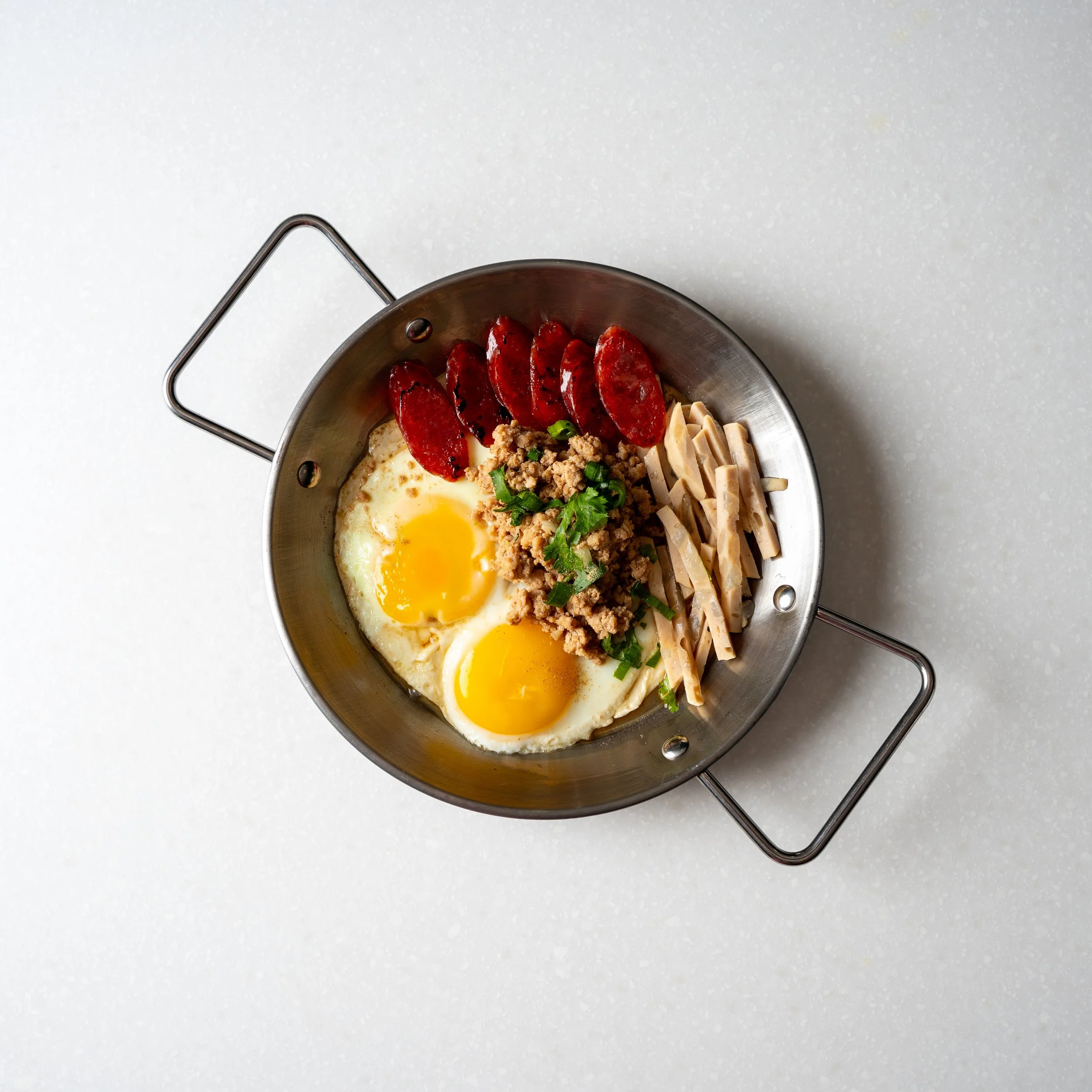 small personal sized metal skillet containing two eggs, sliced sausage meats, strips of pork, ground pork and cilantro garnish