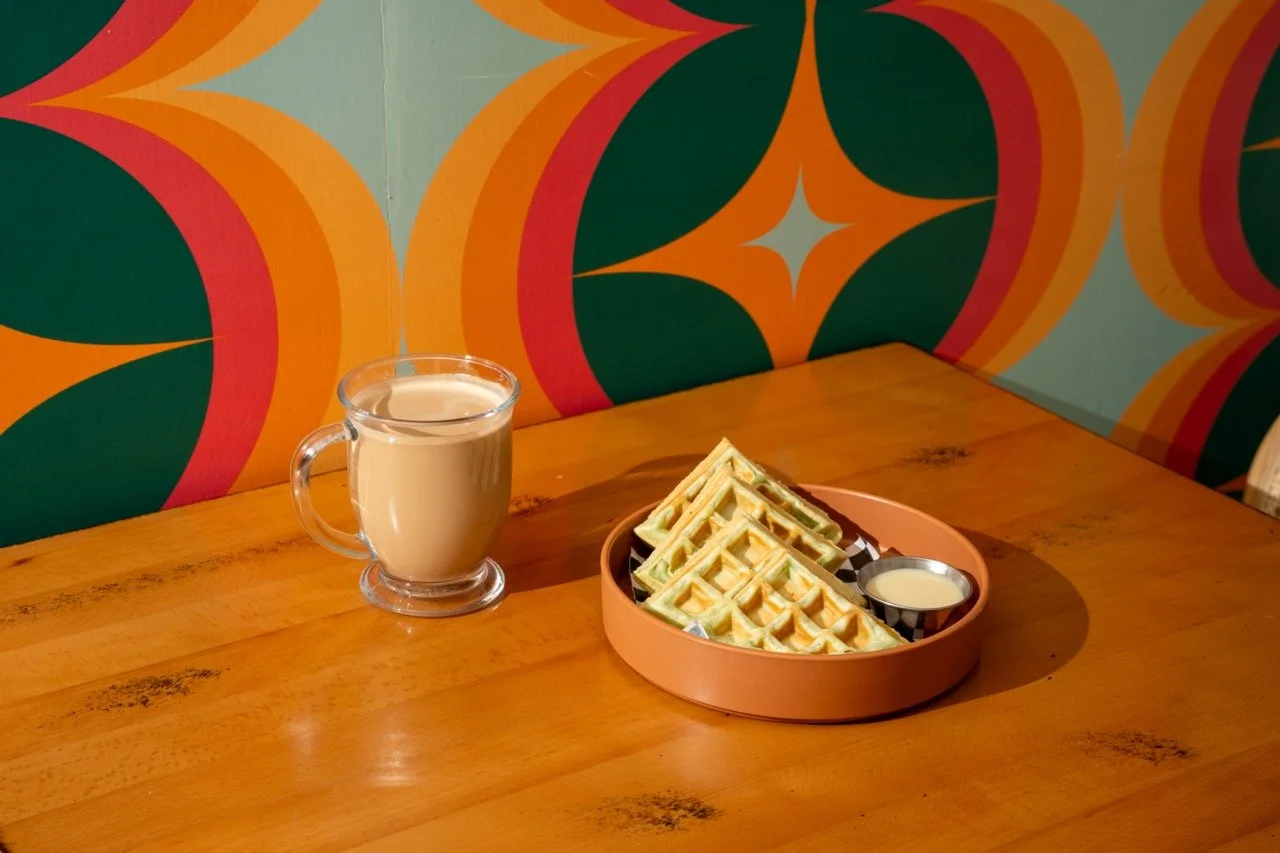 Table with a clear glass mug filled with a hot latte and an orange walled plate with 4 pandan waffle slices and a small dipping container with condensed milk. Background is a colorful retro-patterned wallpaper giving a 1970s aesthetic.