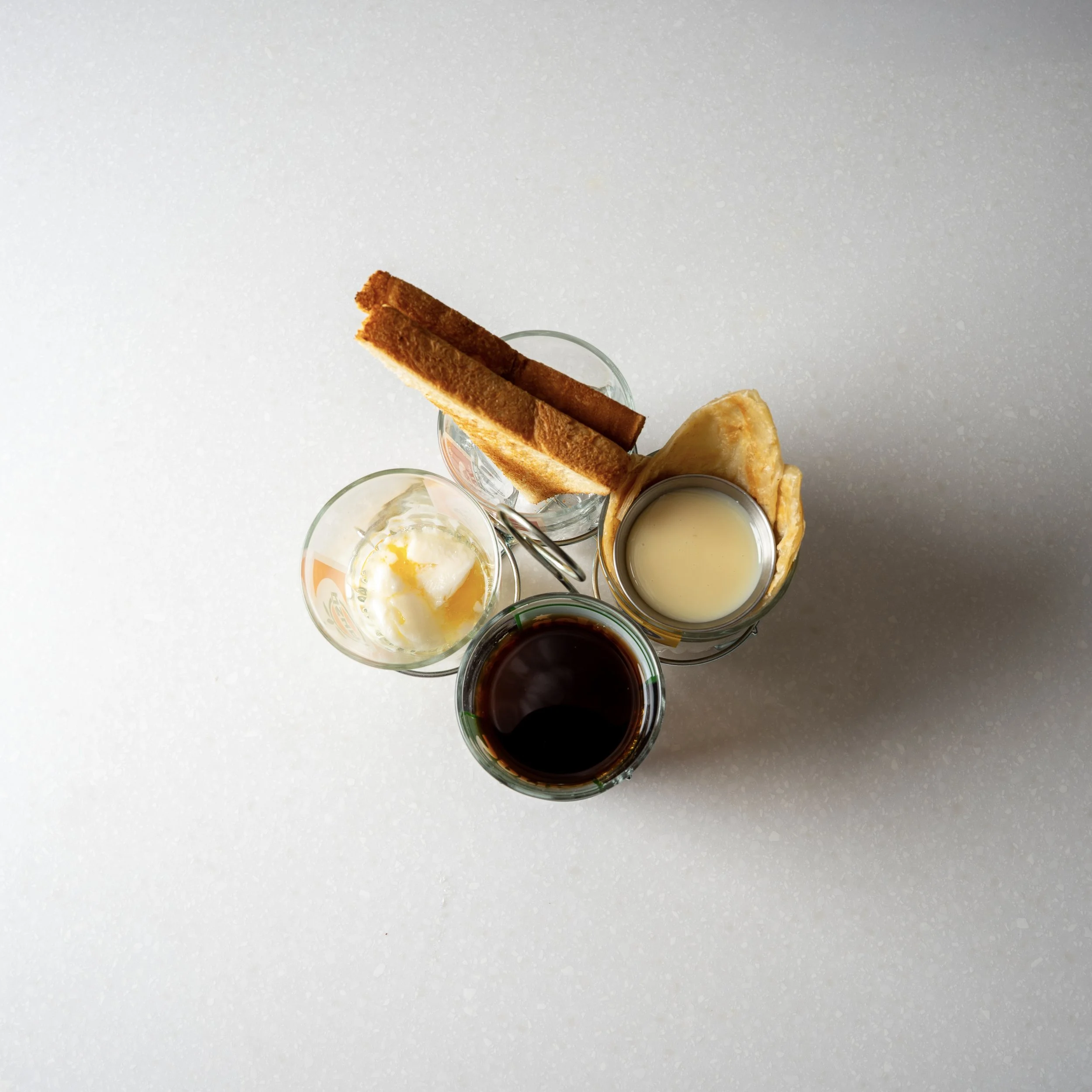 top down view of 4 clear cups, each filled with one of the following:  two soft-boiled eggs, black coffee, roti with condensed milk, crispy toast