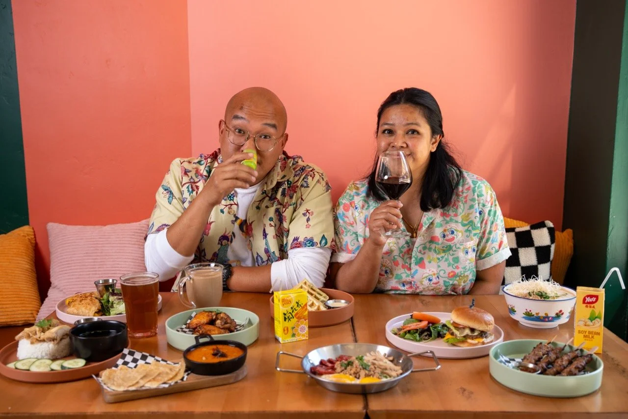 bald man holds Asian nasal inhaler and black-haired women holds glass of red wine up to nose. Both are seated behind a table filled with different plates of food and mugs of coffee and cartons of classic asian beverages
