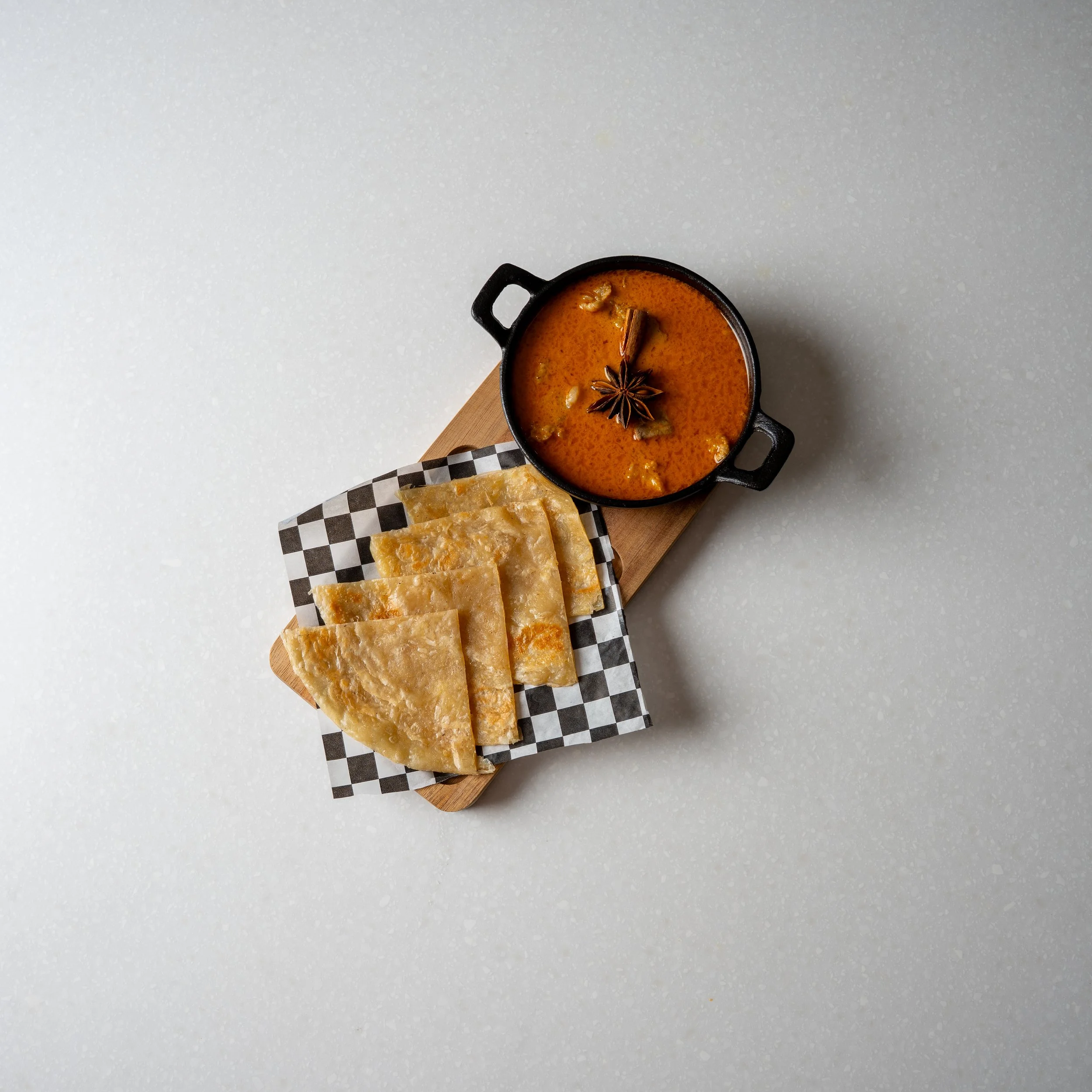 wooden board with black and white checkered deli paper toped with crispy roti slices and a small cast iron skillet filled with curry for dipping