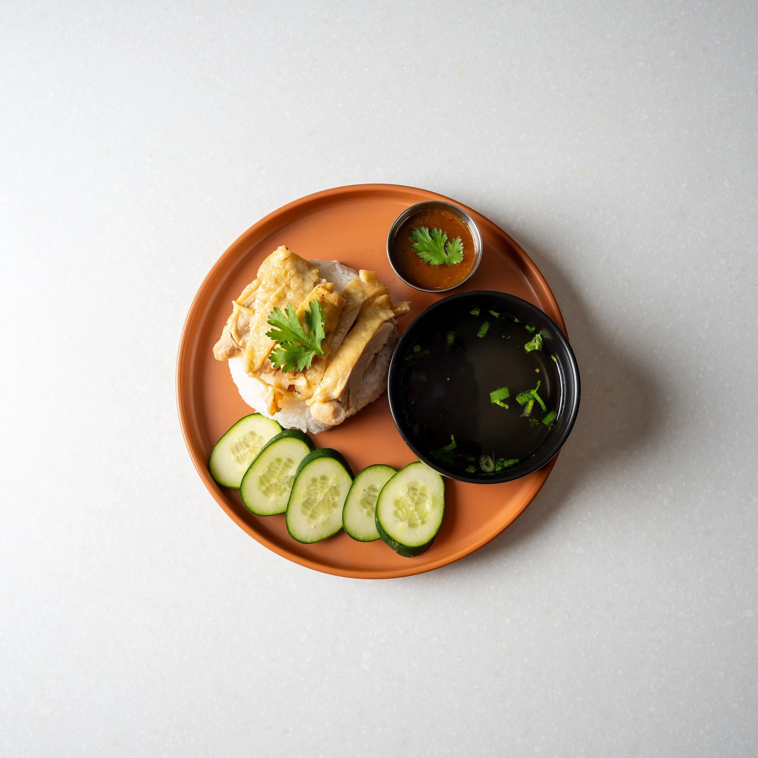 orange plate with rice topped with chicken slices, bowl of soup broth, side of sauce, and sliced fresh cucumbers