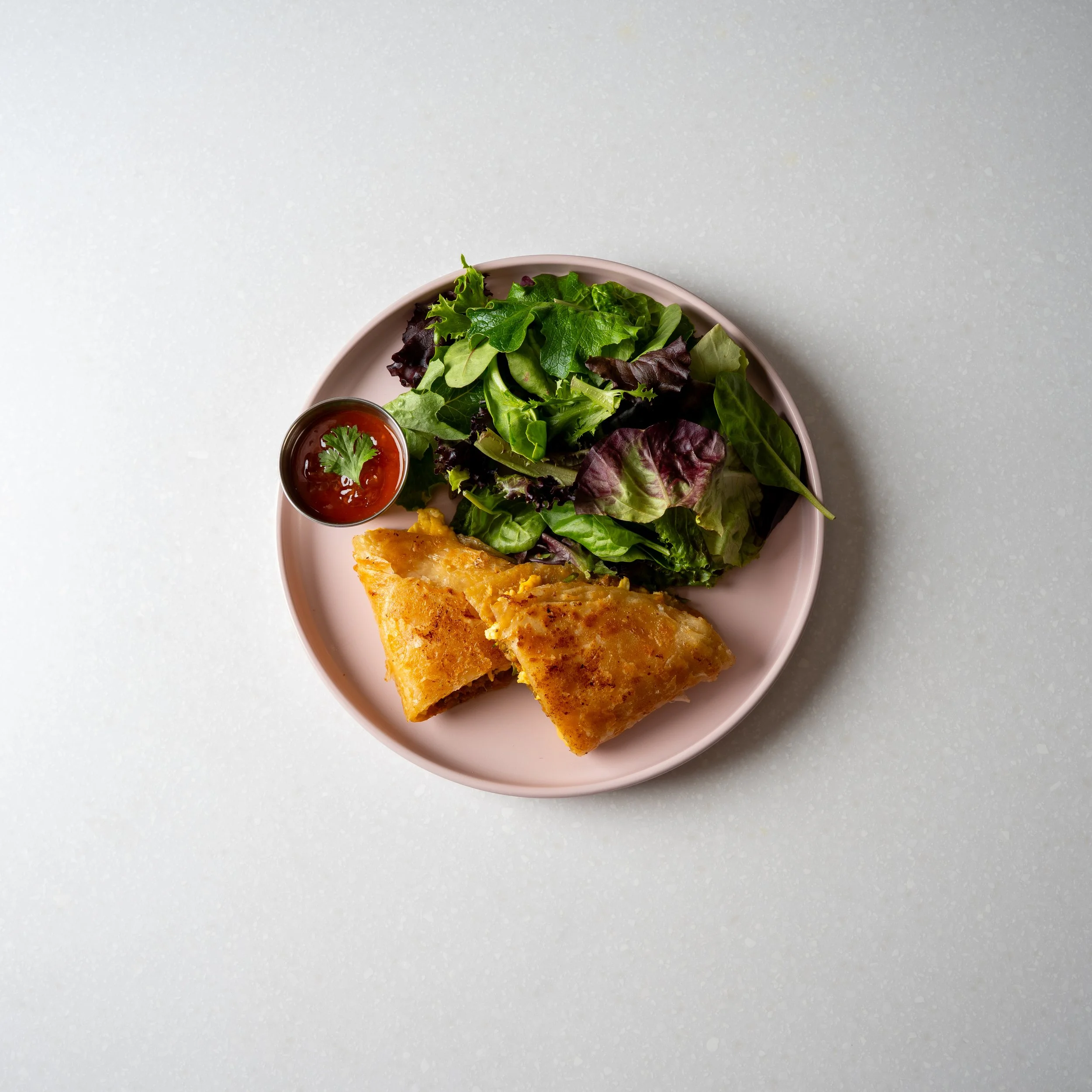pink plate contains two roti sandwiches, a side salad and a reddish chili jam on the side