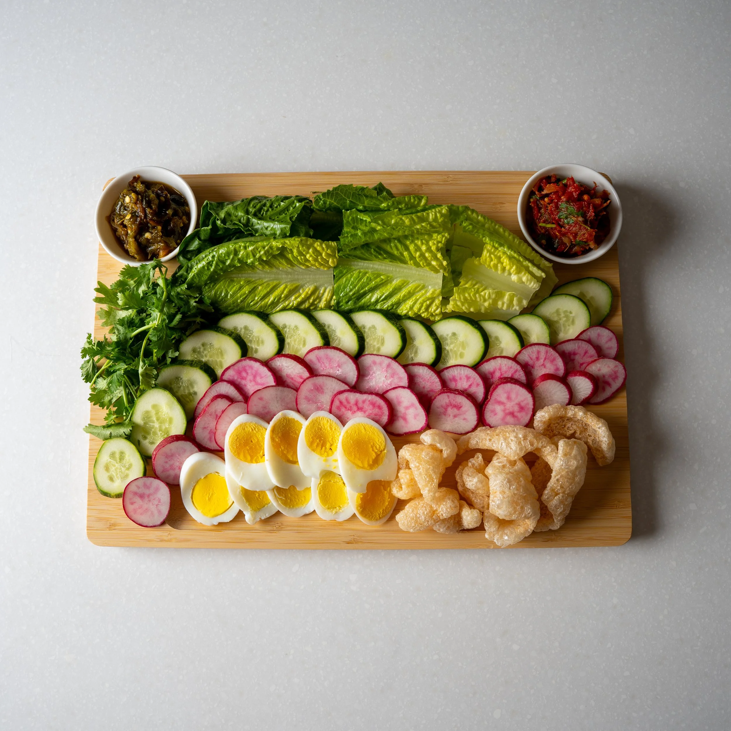 large wooden board filled with sliced vegetables, eggs and pork rinds, very colorful and beautiful, and two sauces for dipping