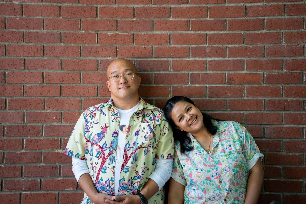 bald man with glasses wearing an ethnic-printed collared shirt leans against brick wall, mid-length haired women also wearing a patterned collared shirt smiles with her head leaning on man's shoulder