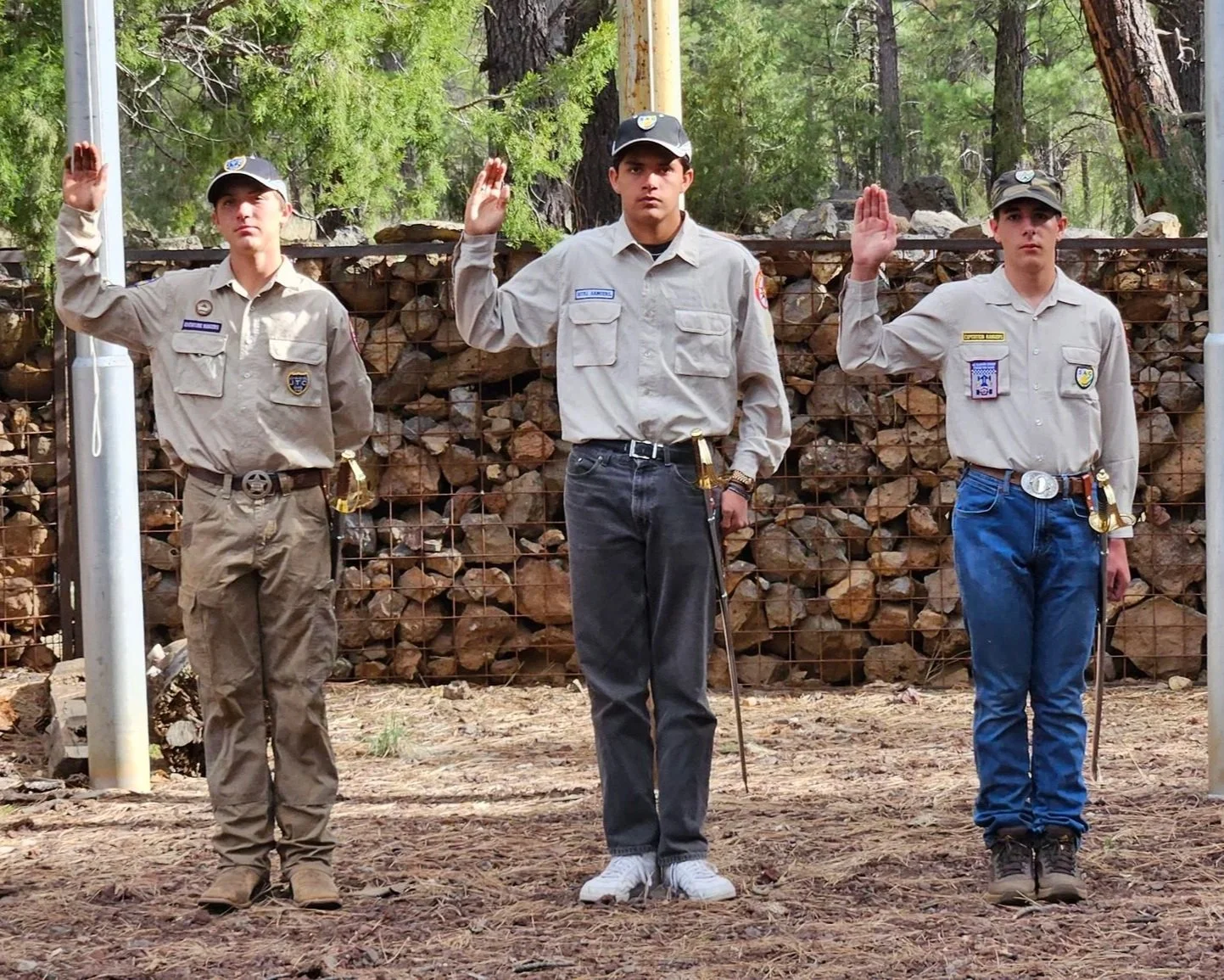 Three young boys wearing Royal Ranger uniforms with badges and sashes, standing outdoors in front of a wire fencing filled with rocks, with trees visible in the background. They are raising their right hands in a salute.