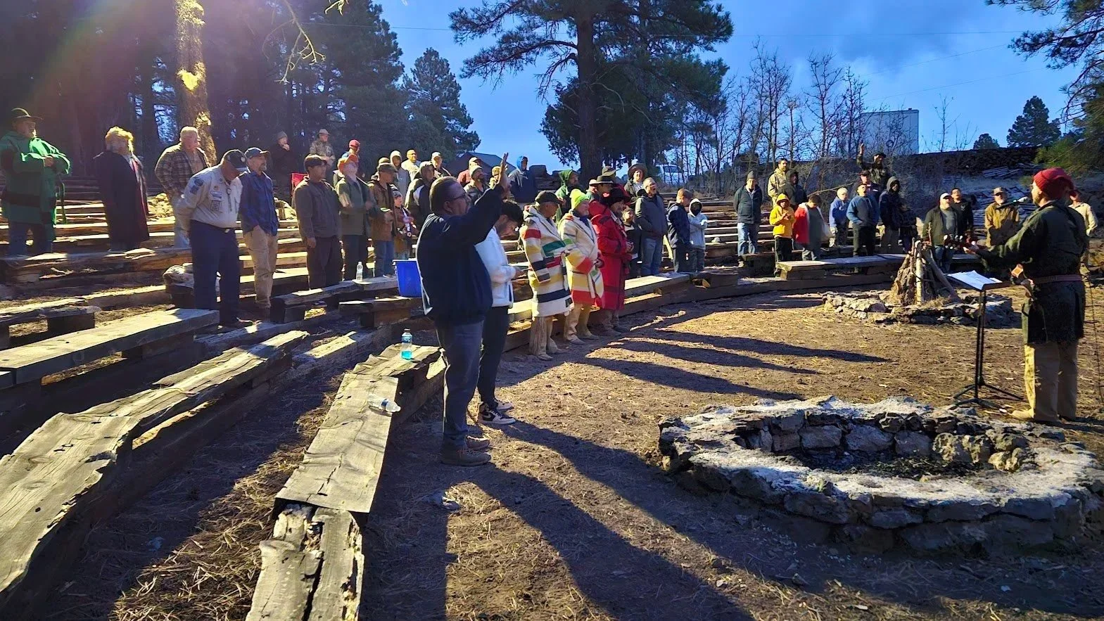 Group of Royal Rangers standing outdoors on tiered wooden benches during a ceremony or gathering, with a speaker at a microphone near a fire pit, in a wooded area with tall trees and a clear blue sky.