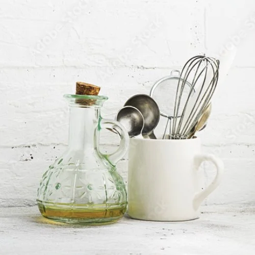 A glass oil bottle with a cork, a white mug holding kitchen utensils including a whisk, a measuring cup, and a ladle, set against a white brick wall background.