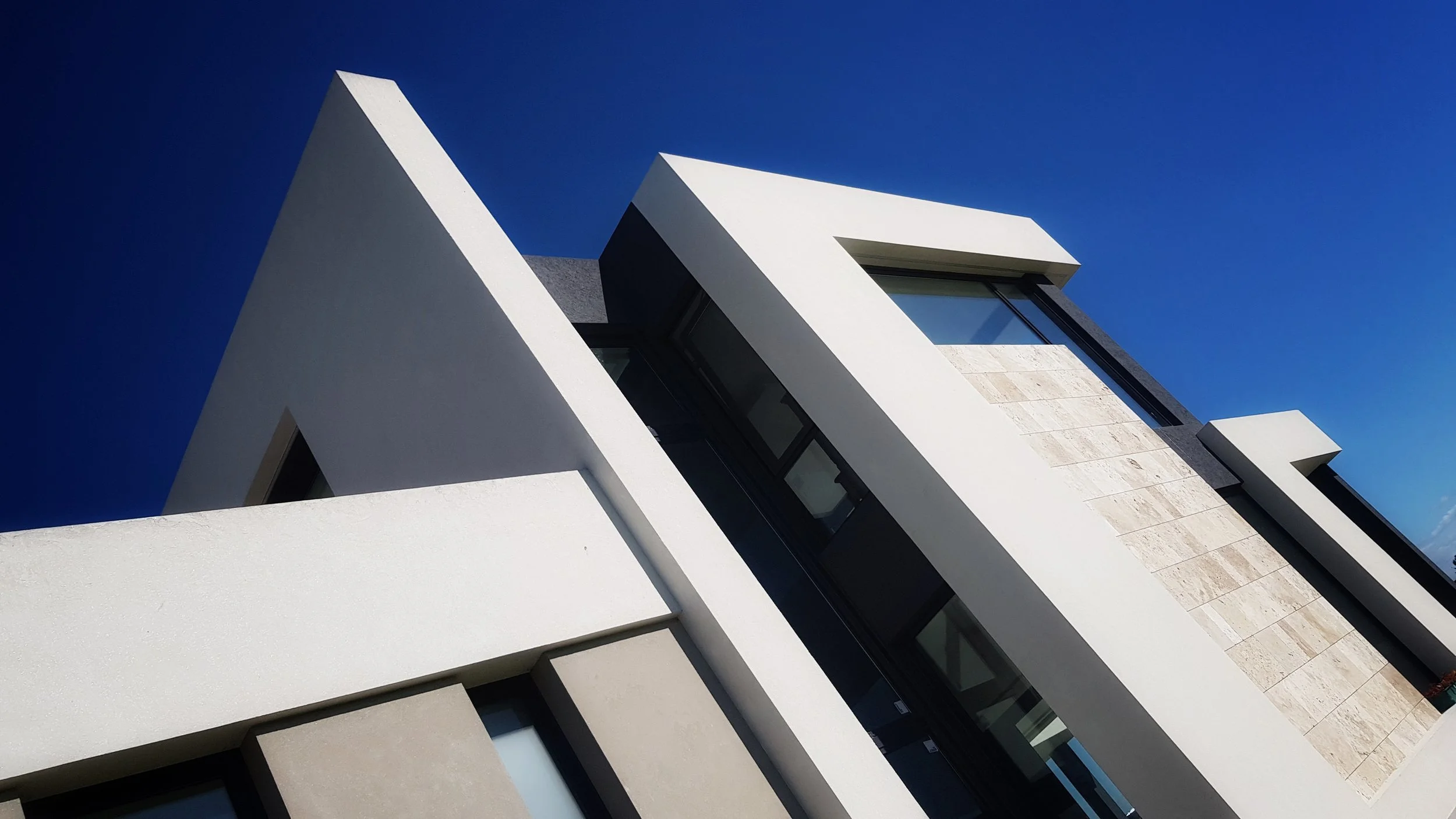 Vista frontal de un edificio moderno con formas geométricas, paredes blancas y detalles en beige, con ventanas grandes y un cielo azul de fondo.