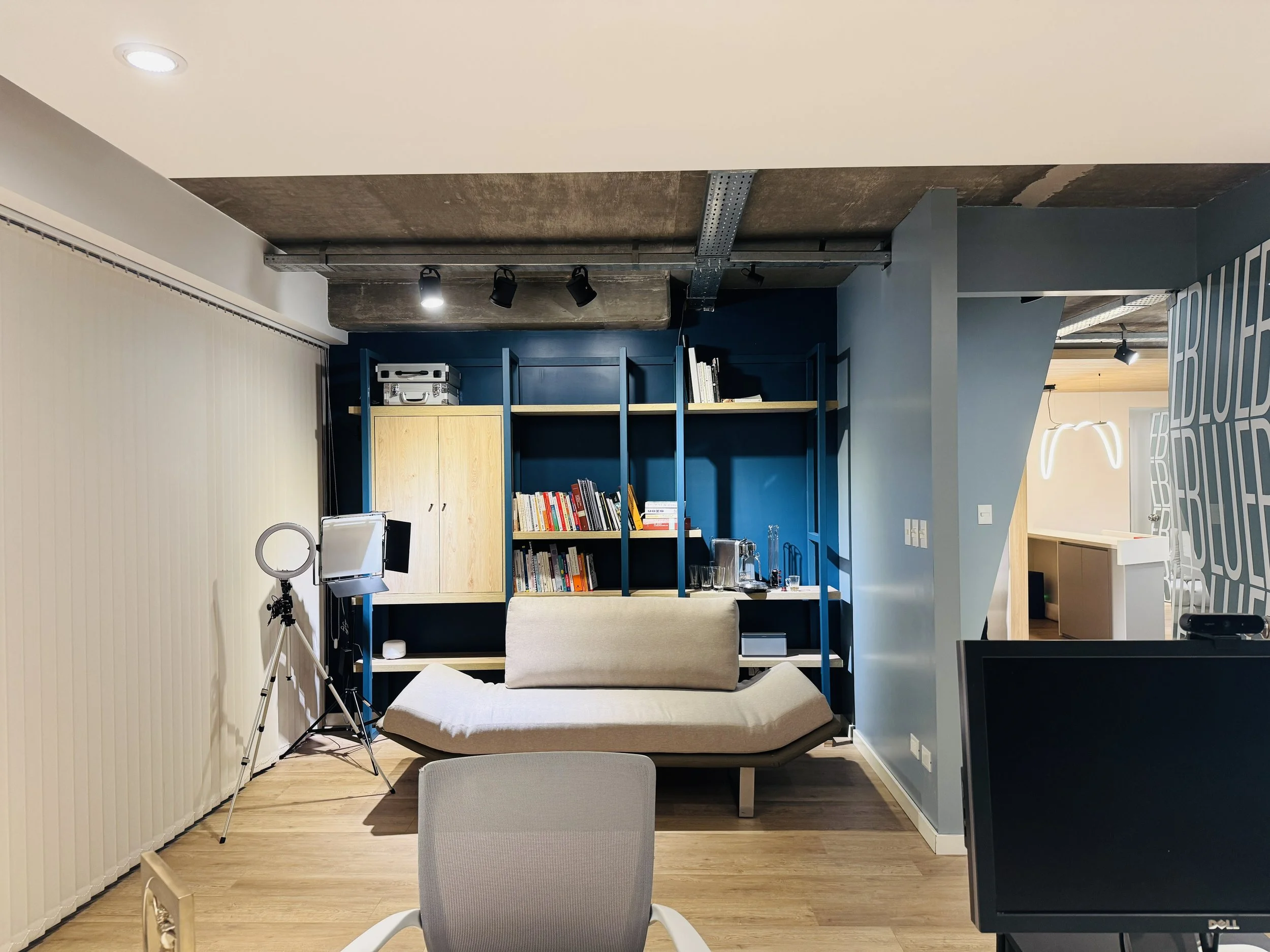 Sala moderna con estantería azul y madera, sofá beige, equipo de grabación, y monitor en un espacio de oficina o estudio.