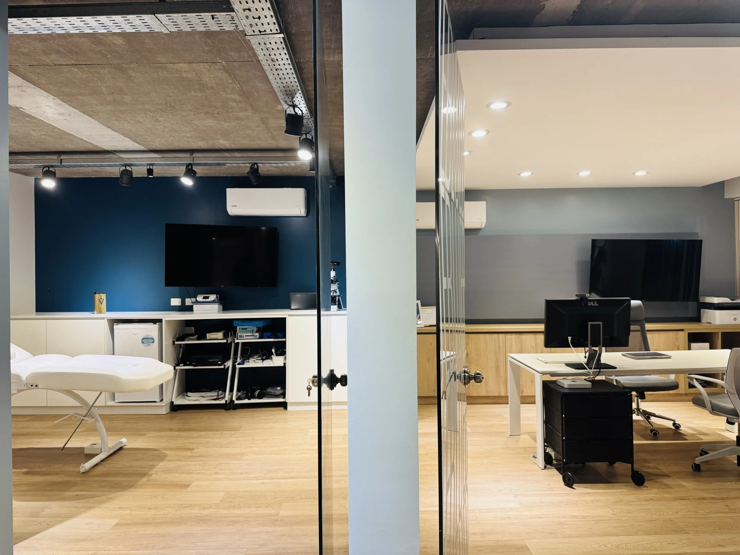 Oficina moderna con dos áreas separadas por una pared. La izquierda tiene una pared azul con televisión, escritorio y equipo de oficina, y un banco de examen médico. La derecha tiene paredes grises, oficina con escritorios, computadoras y mobiliario 