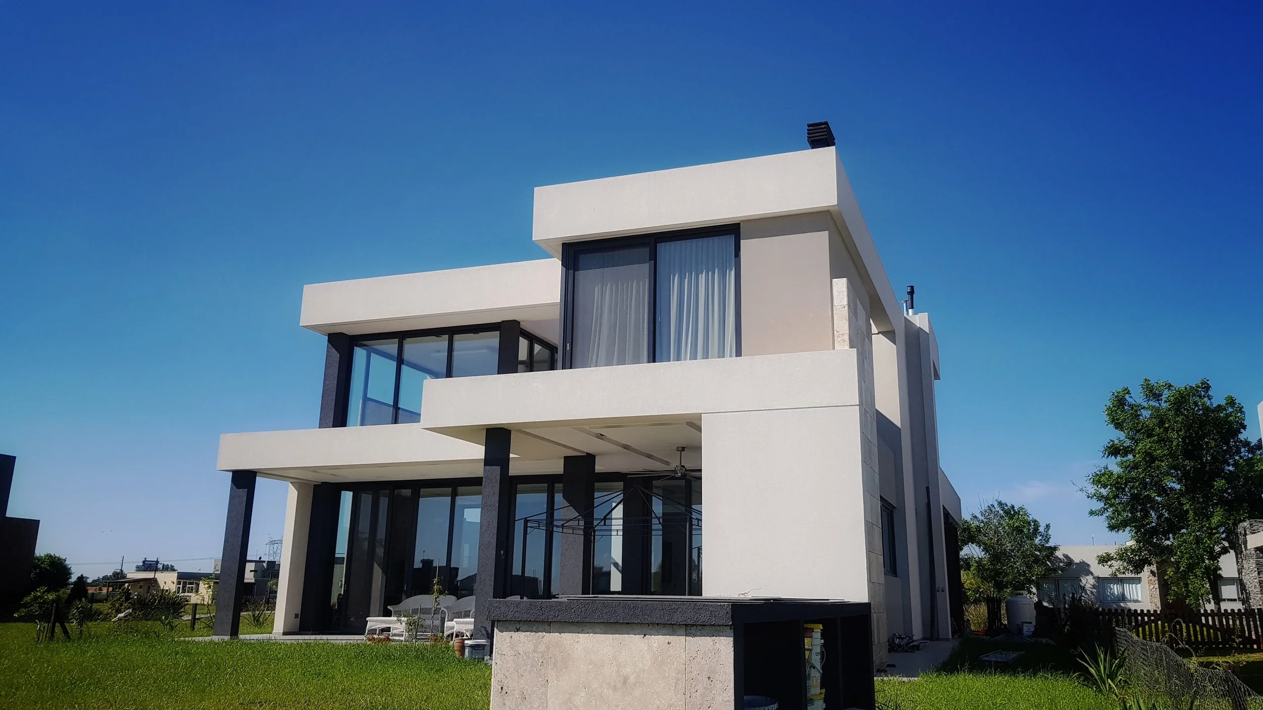 Casa moderna de tres pisos con enormes ventanas de vidrio, estructura de color blanco y negro, en un día soleado con cielo azul y césped verde.