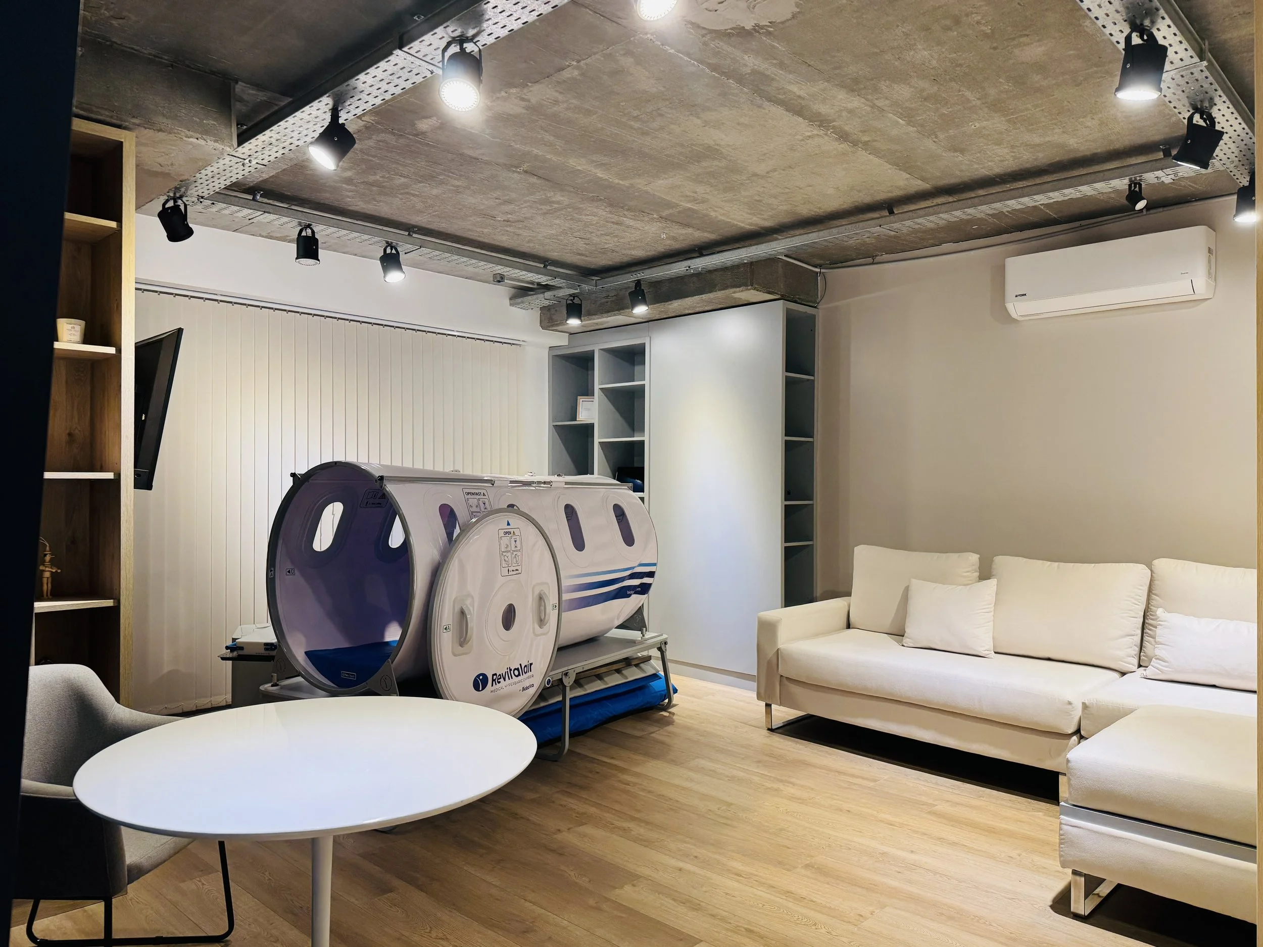 Habitación de con sala de estar y un cápsula de tratamiento, sillón, mesa circular, estantería con libros y una pared con cortina. El techo de concreto tiene luces empotradas y rastreles con luces dirigidas.